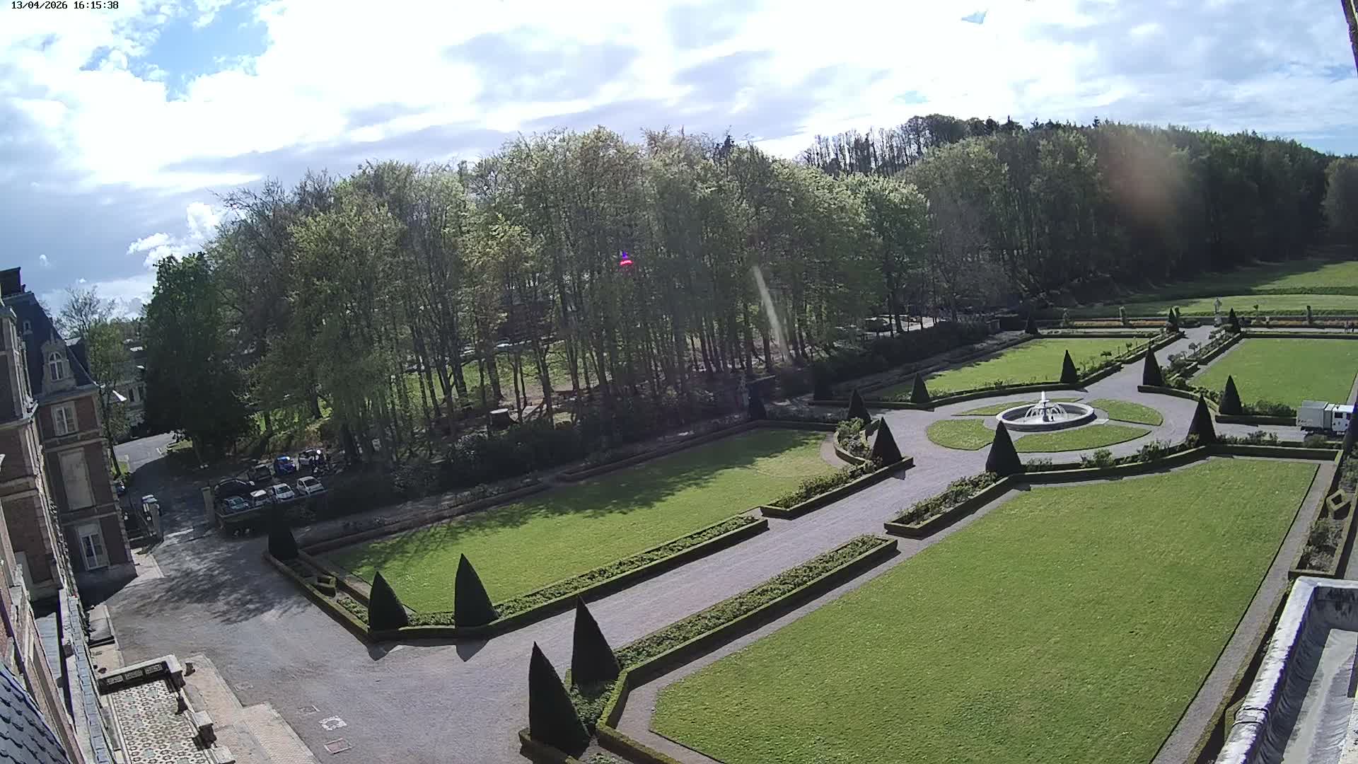 An overcast day reveals a formal garden with a central fountain, manicured lawns, and hedges, adjacent to a large building.