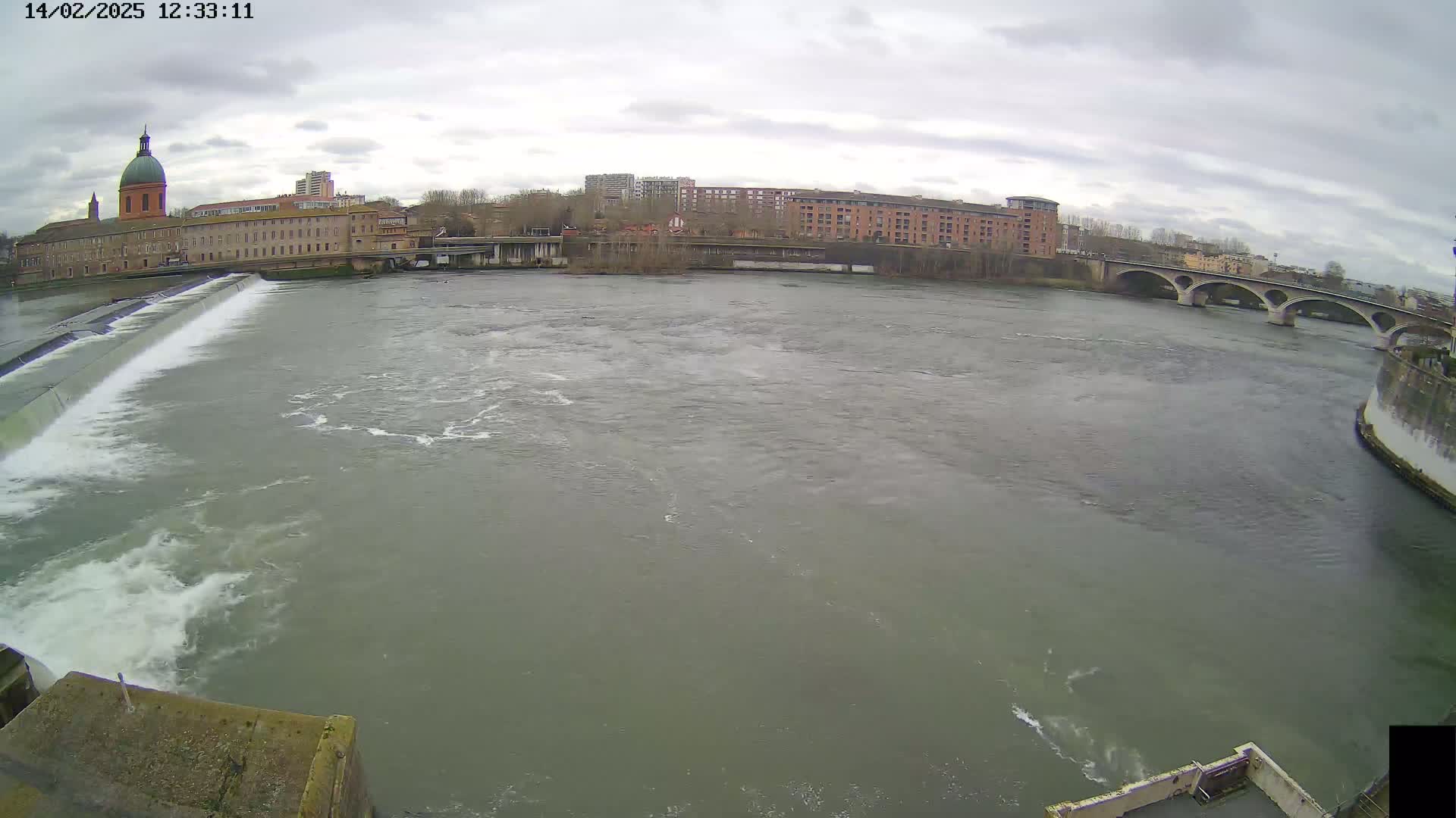 A wide shot of a river flowing past a dam and under a bridge on an overcast day, with buildings and trees visible on the opposite bank.
