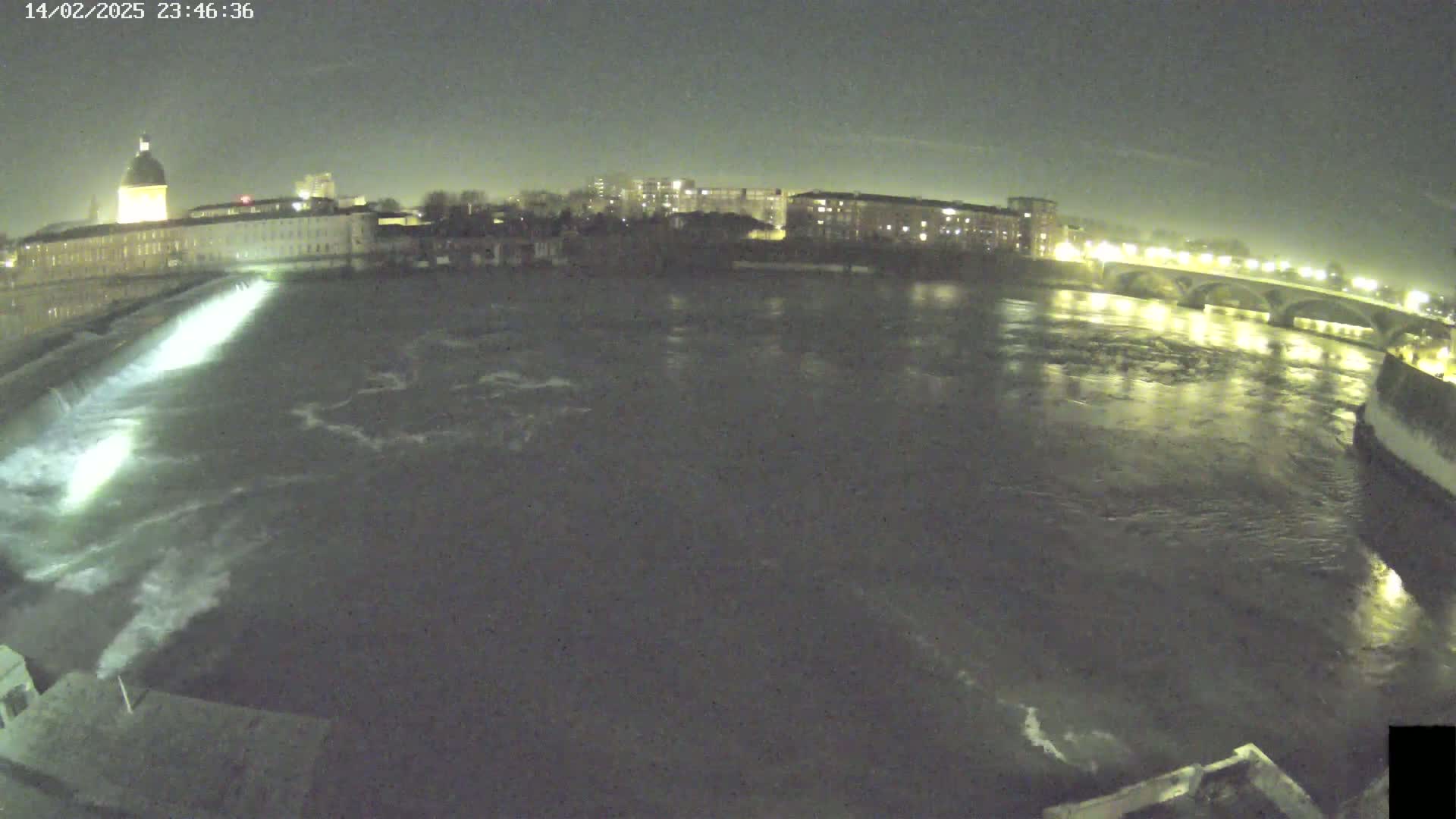A nighttime view of a river flowing past city buildings and a bridge under a dark sky.