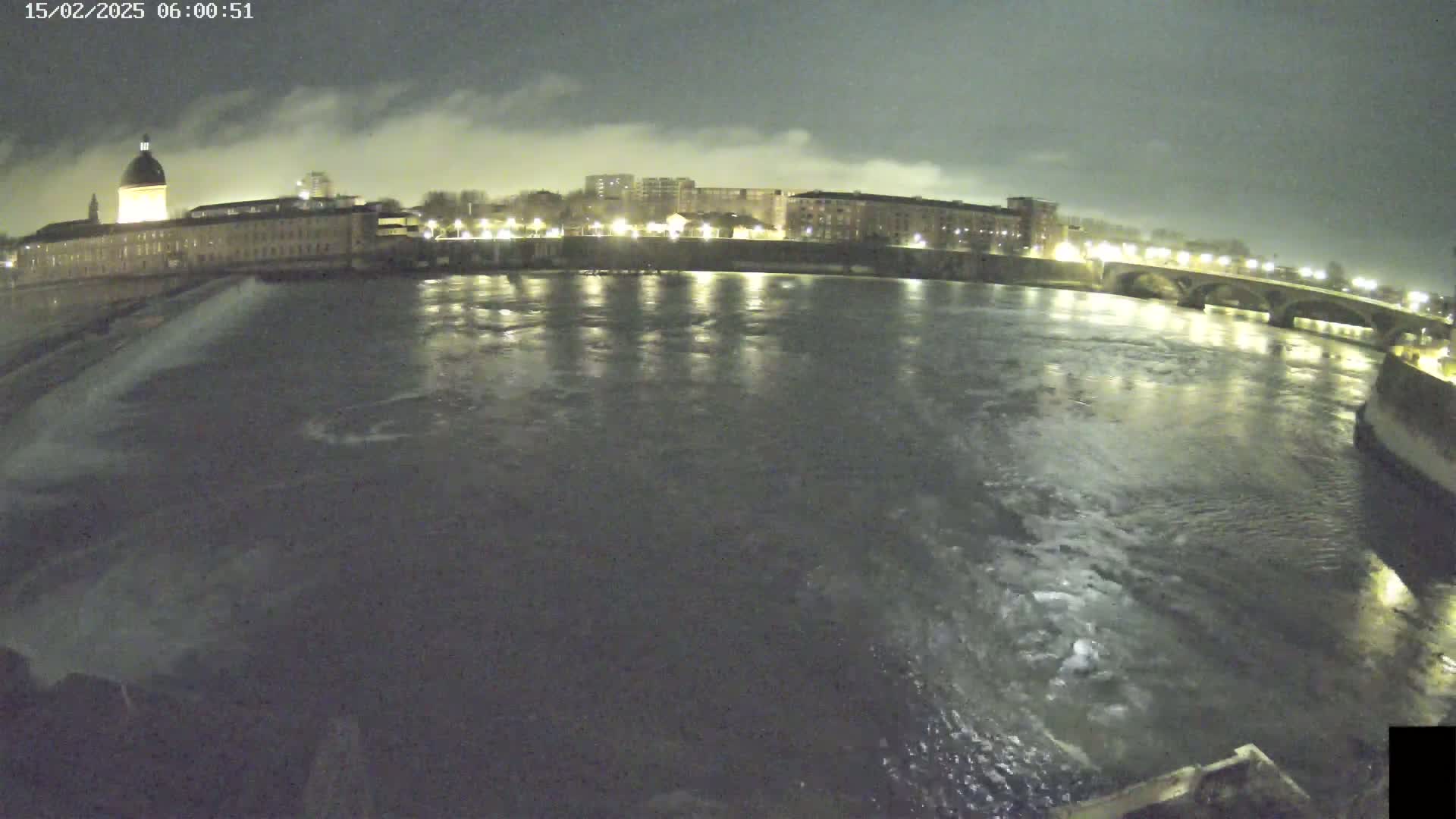 A wide nighttime shot shows a river flowing past a city with buildings lit up under a cloudy sky.