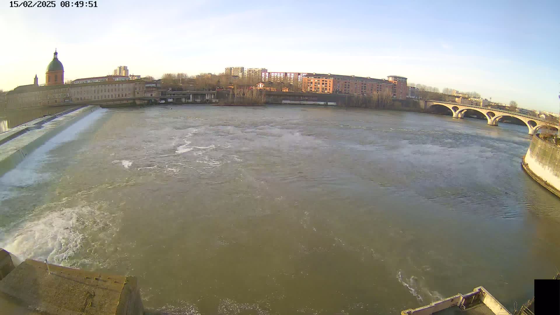 A wide shot of a river flowing past a dam and under a bridge, with buildings and a domed structure visible in the background under a clear sky.