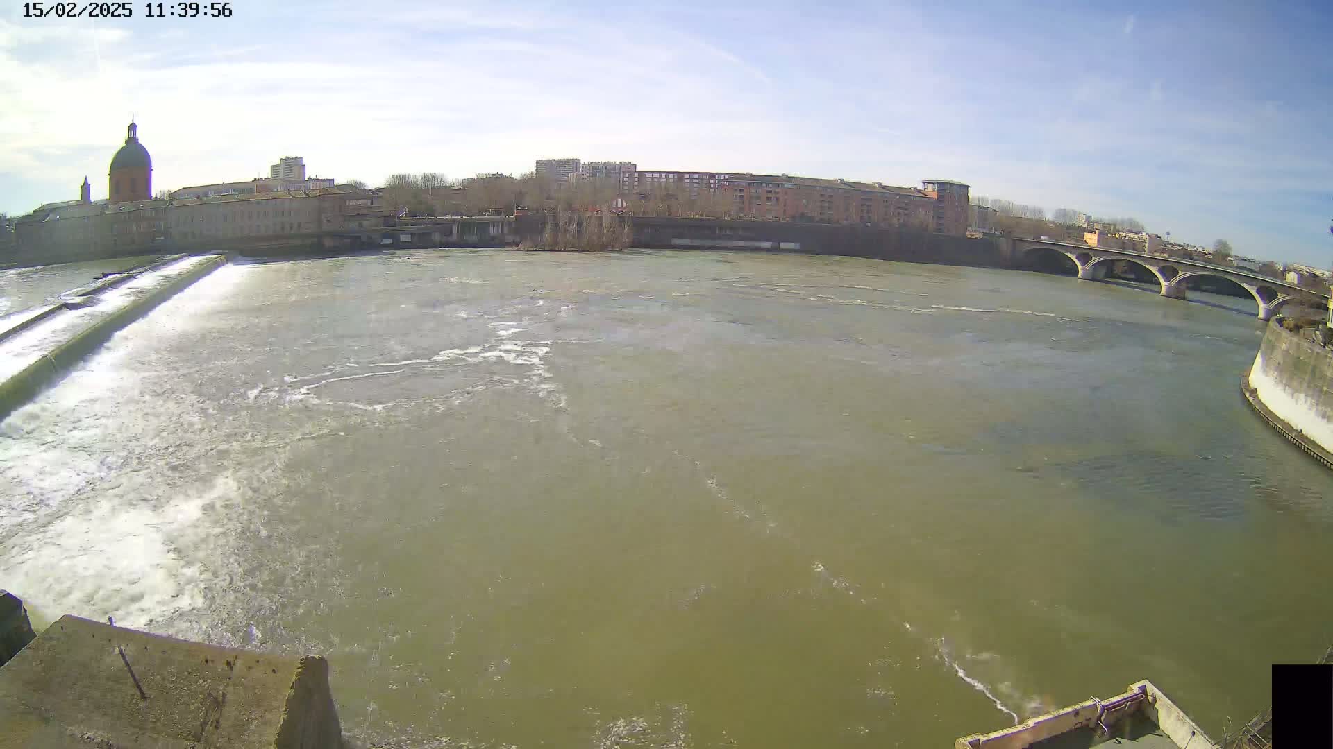 A wide shot of a river flowing past a city under a mostly sunny sky, with a dam visible in the foreground and a bridge in the distance.