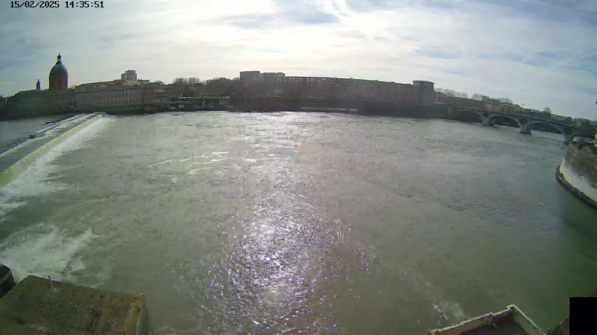 A wide shot shows a river flowing past a city under a partly cloudy sky, with a dam in the foreground and a bridge in the distance.