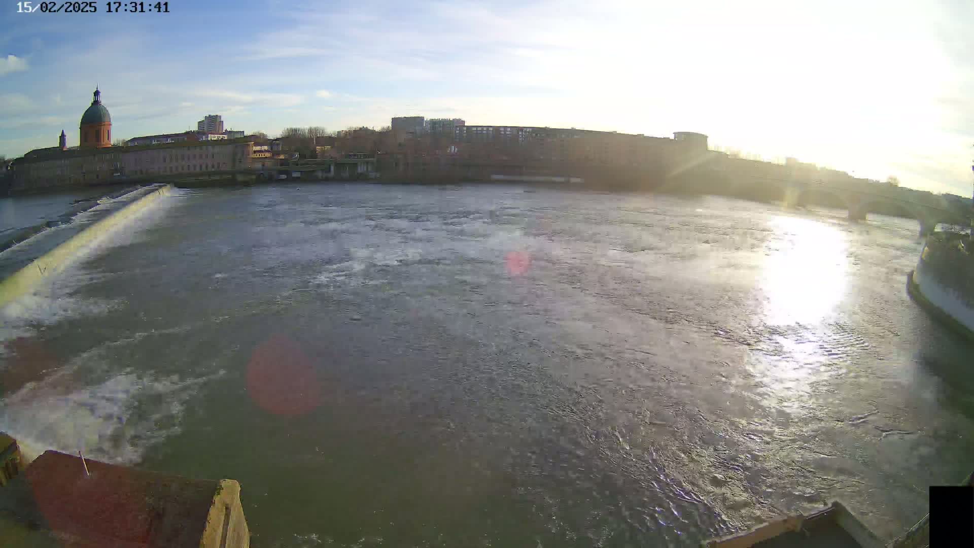 A wide shot shows a river flowing past a city, with a dam visible in the foreground and buildings, including a domed structure, visible across the water under a sunny sky.