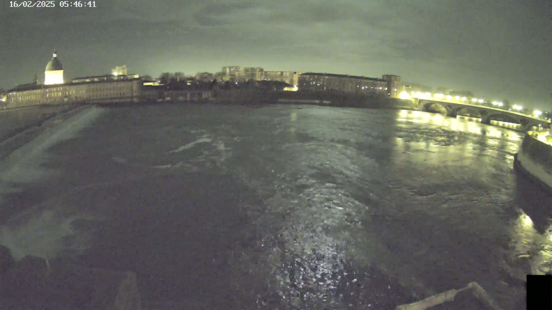 A nighttime view of a river flowing past a city with buildings and a bridge, under a cloudy sky.