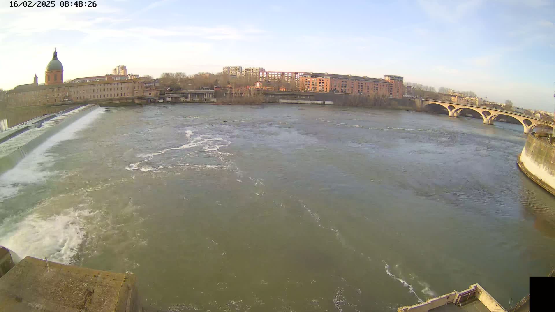 A river flows under a bridge past a dam and buildings under a mostly clear sky.