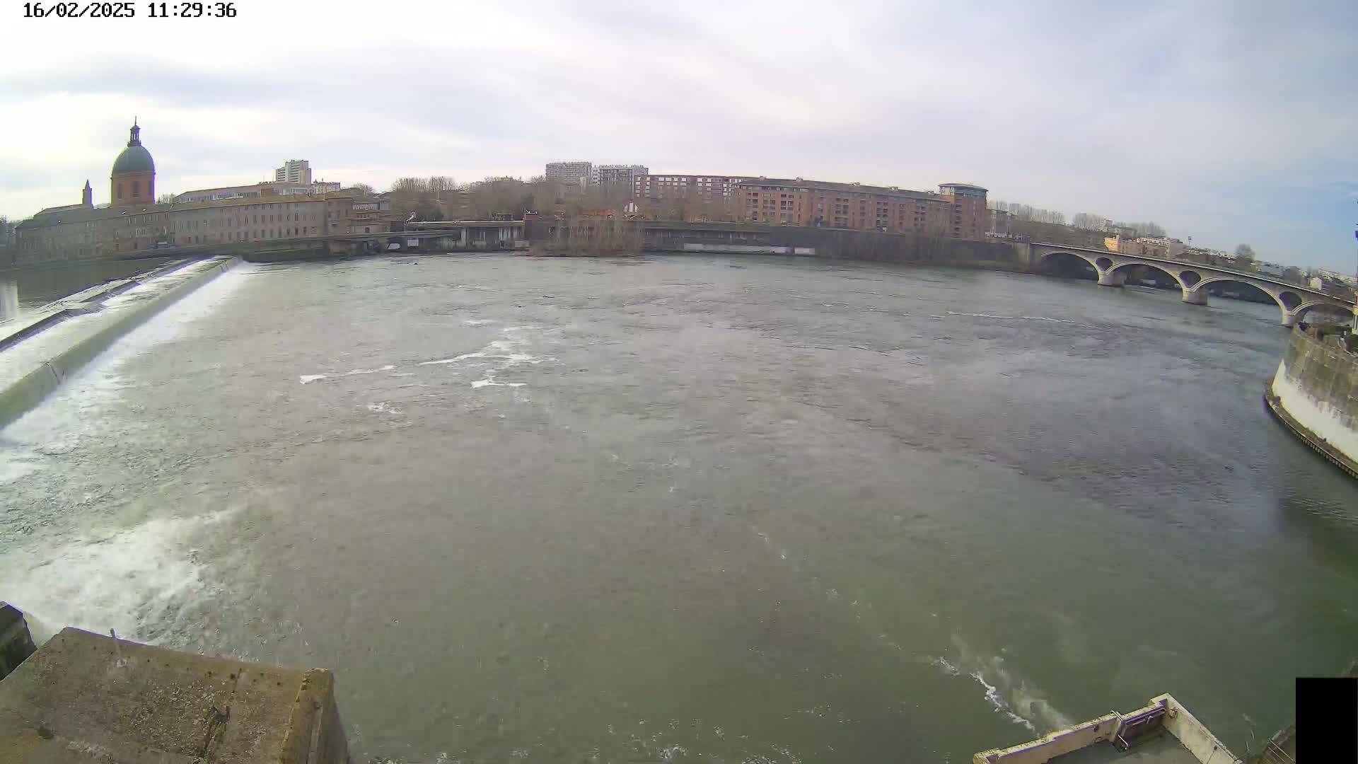 A wide shot shows a river flowing past a dam and under a bridge, with buildings and a domed structure visible on the opposite bank under an overcast sky.