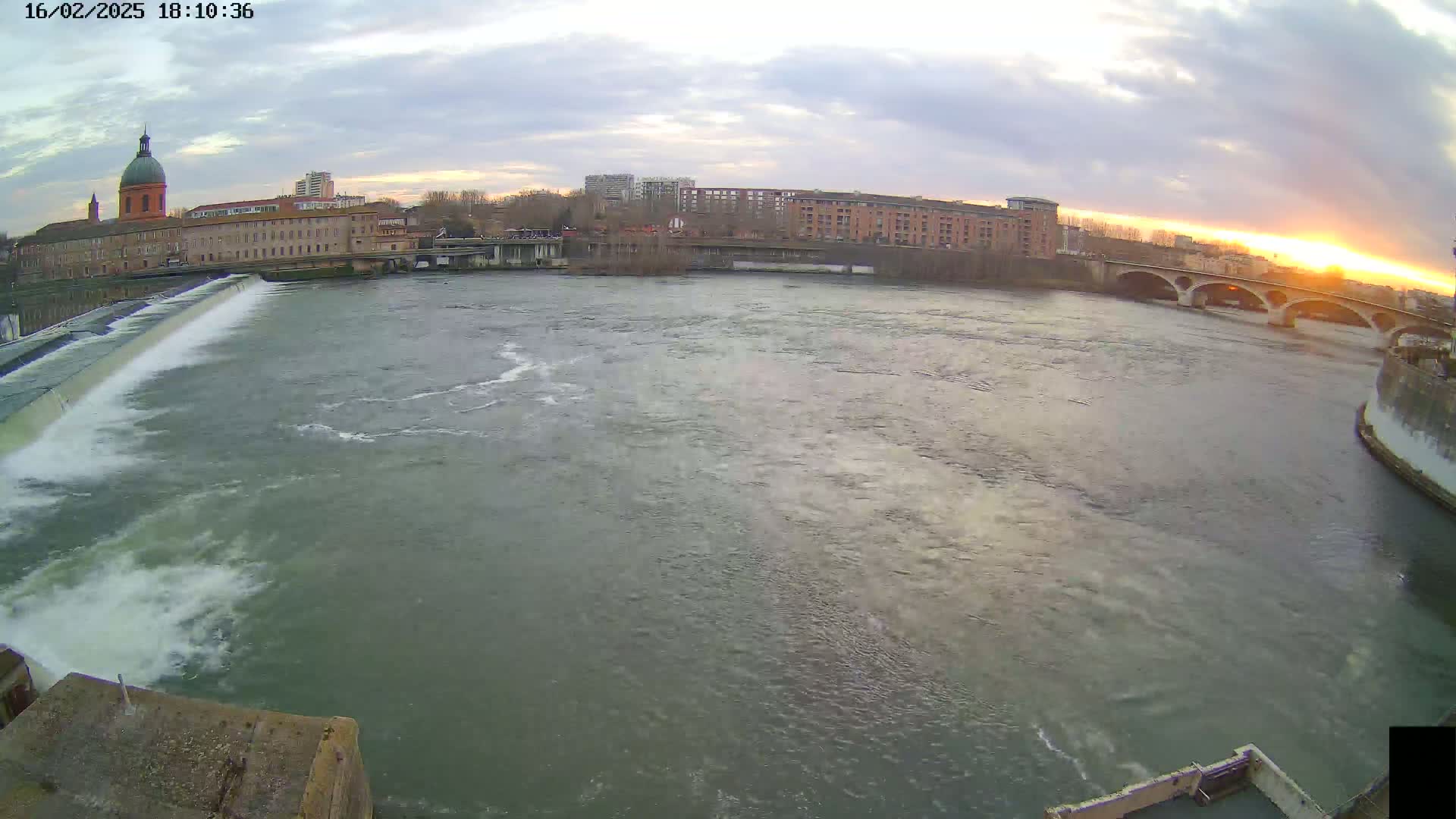 A wide shot of a river flowing past a dam and under a bridge, with buildings and a dome-topped structure visible on the far bank at sunset under cloudy skies.