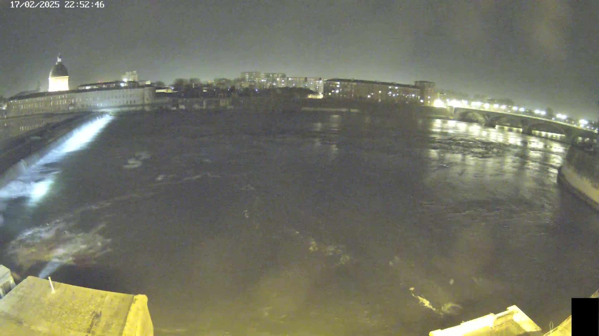 A nighttime view of a river flowing past a city with buildings and a bridge under a dark sky.