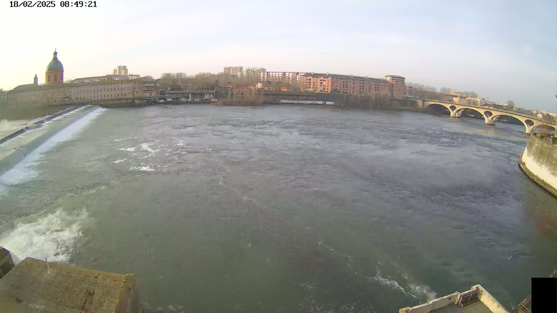 A wide shot of a river flowing past a city, featuring a dam, a bridge, and buildings under a mostly clear sky.