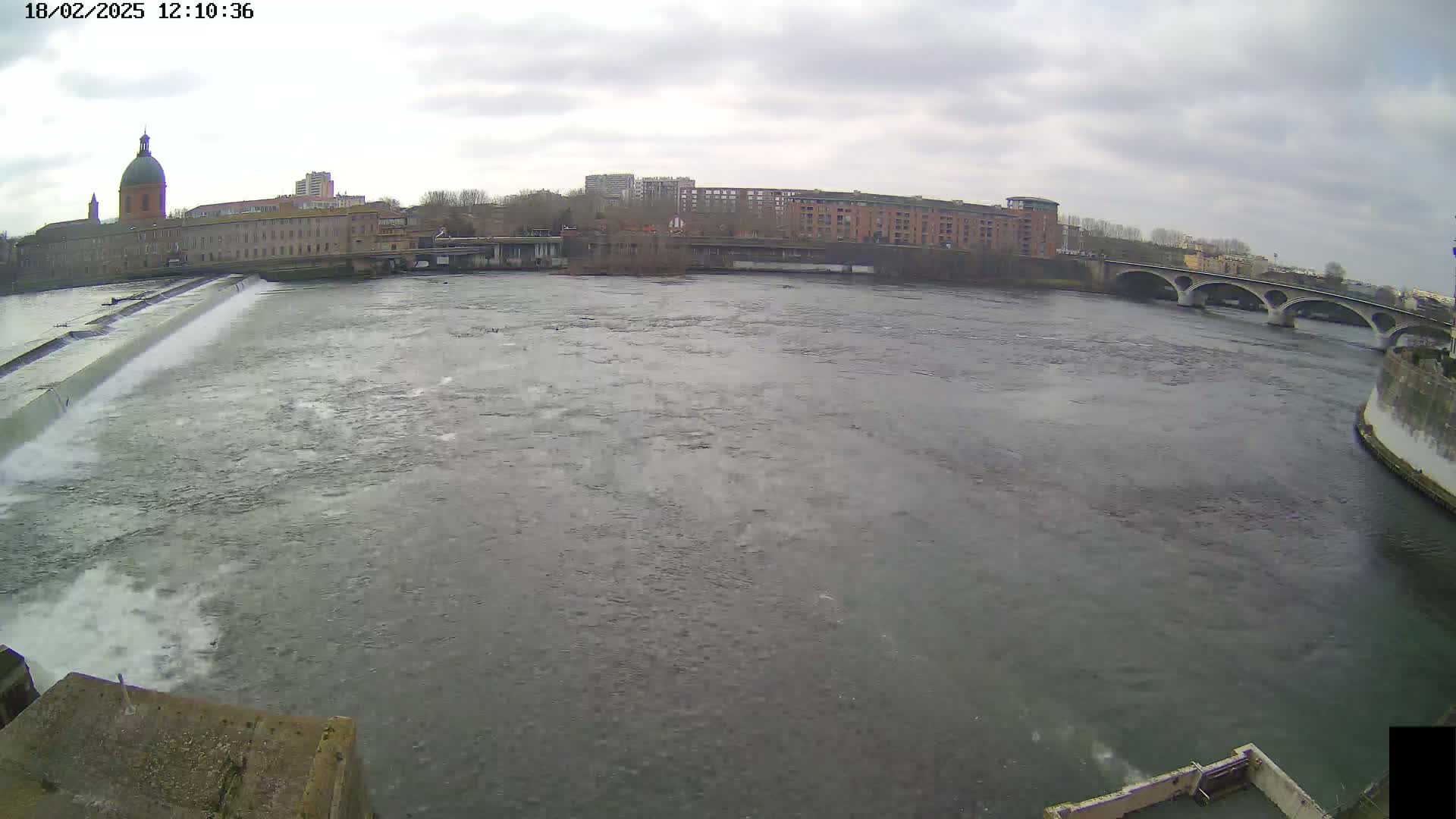 A wide shot of a river flowing past a city under an overcast sky, with a dam and bridge visible in the distance.