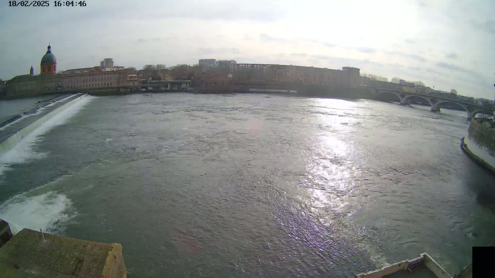 A wide shot of a river with a dam and buildings in the background under a partly sunny sky.