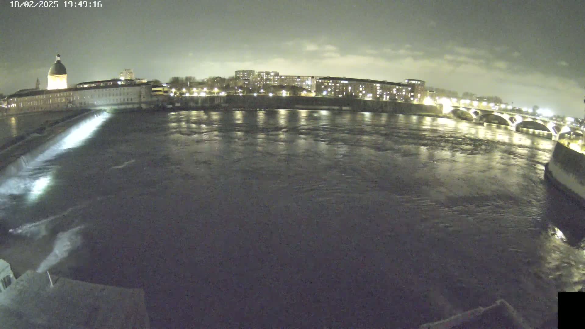 A nighttime view of a river flowing past city buildings and a bridge under a cloudy sky.