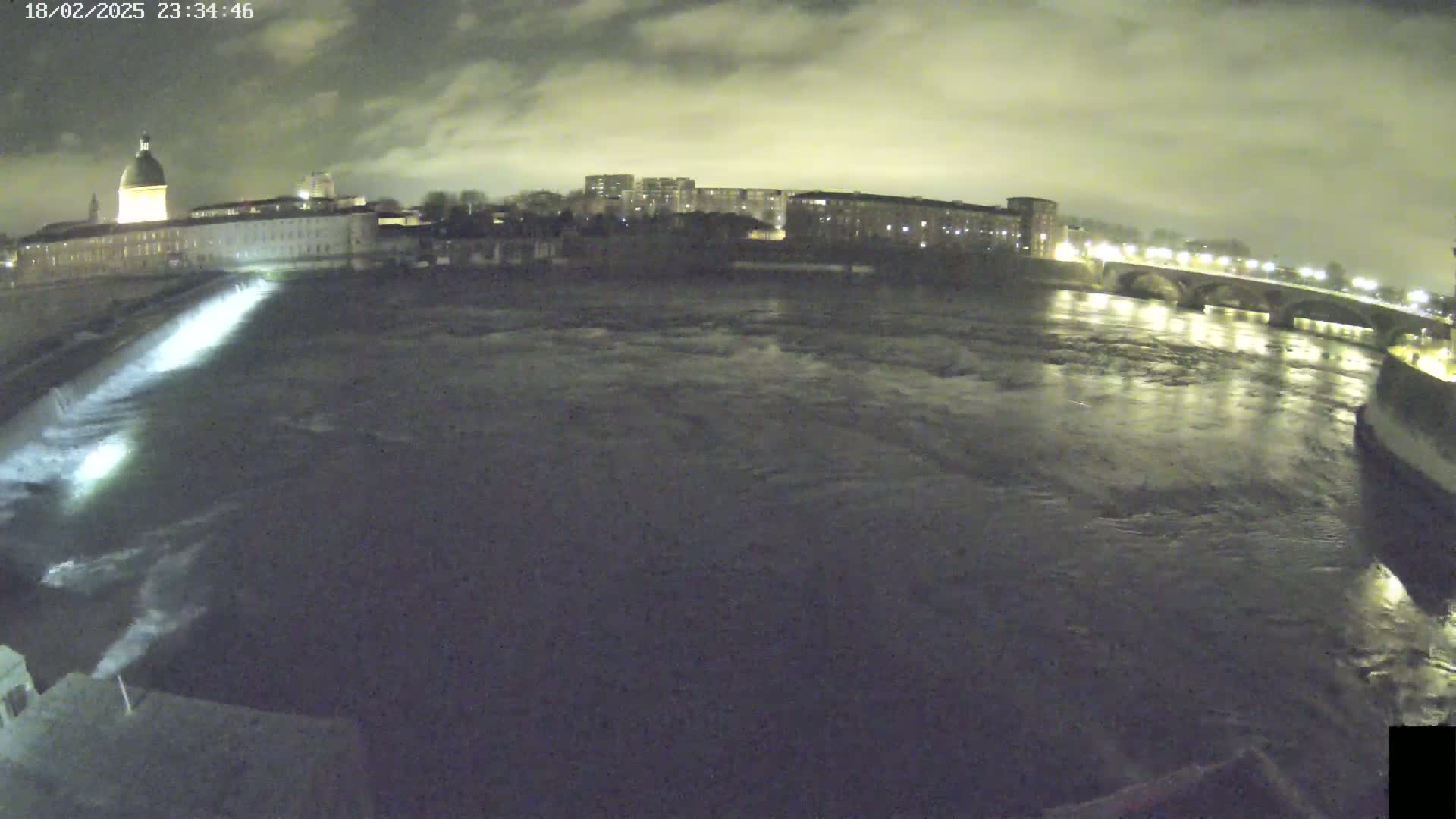A nighttime view of a river flowing past a city, under a cloudy sky, with a dam and bridge visible.