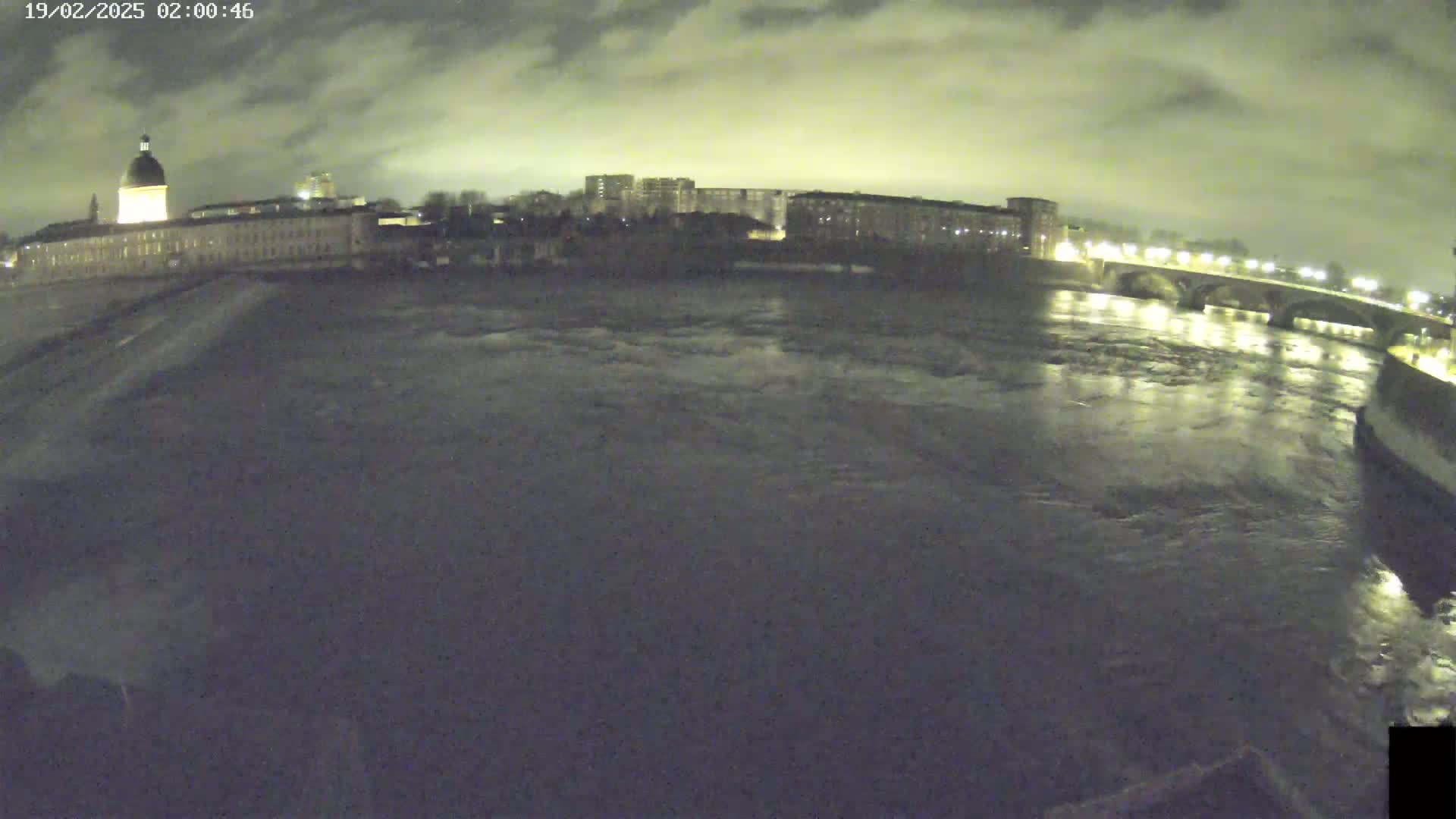 A nighttime view of a river flowing under a bridge, with buildings and lights visible on either side under a cloudy sky.