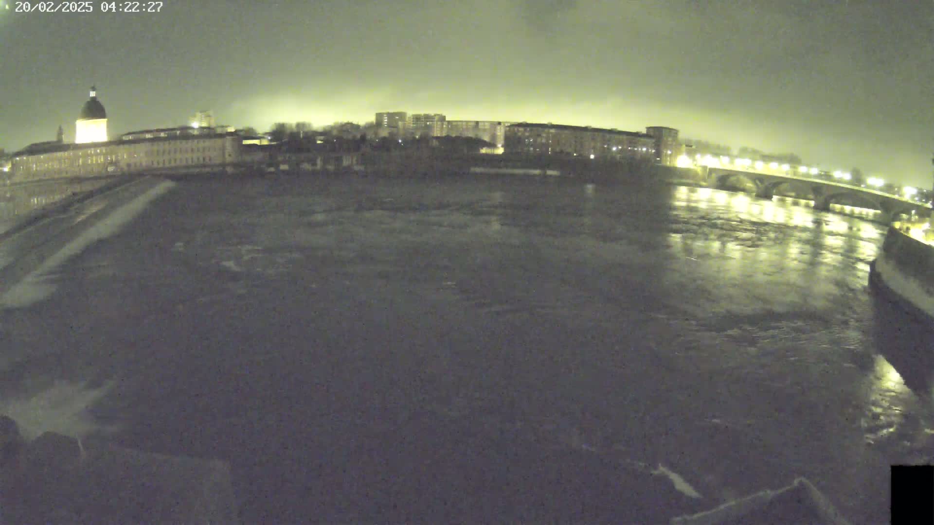 A dark, nighttime view of a river flowing past a city with buildings and a bridge under a cloudy sky.