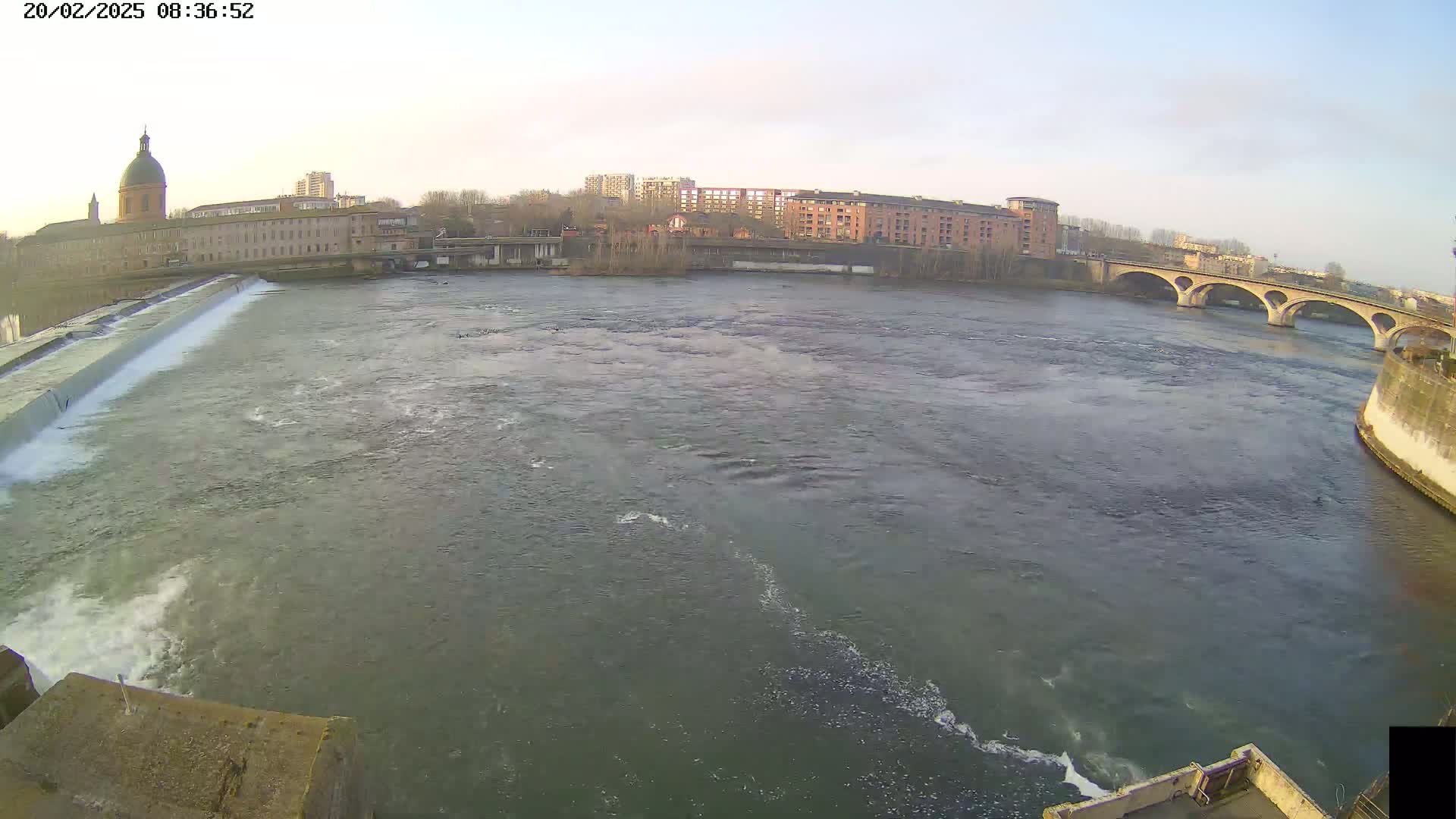 A wide river flows under a stone bridge past buildings and a dam, under a mostly clear sky.