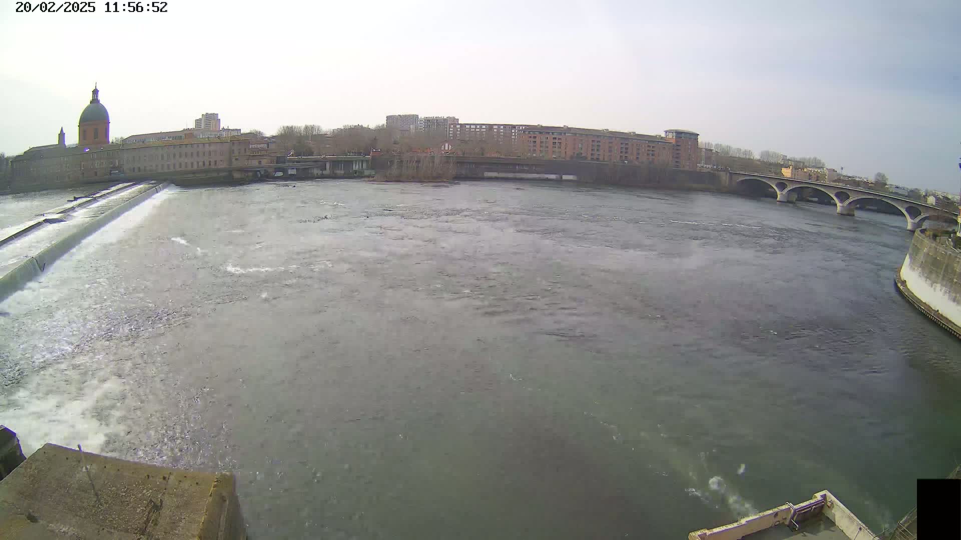 A wide shot shows a river flowing past a dam and under a bridge, with buildings and a domed structure visible in the background under an overcast sky.