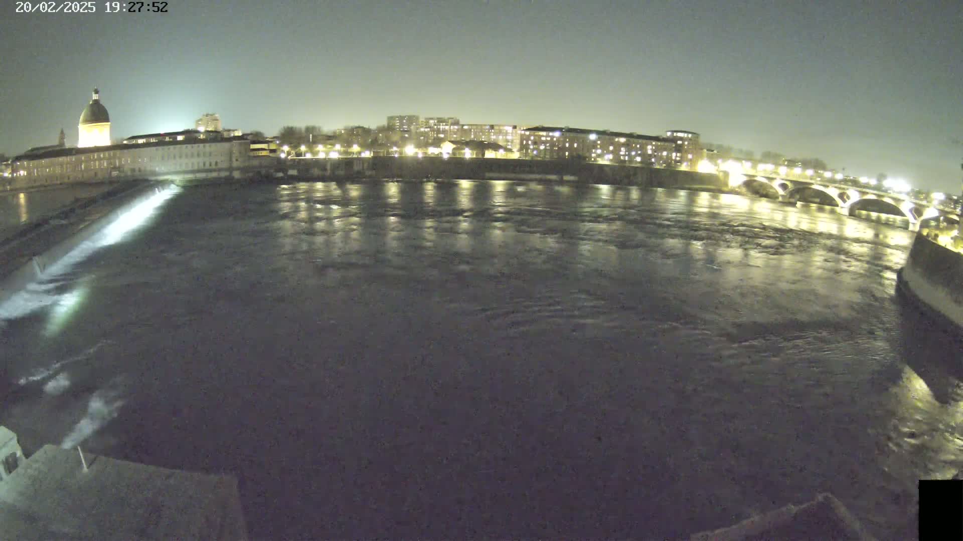 A nighttime view of a river flowing past a city, with a dam visible in the foreground and a lit bridge in the background under a dark sky.