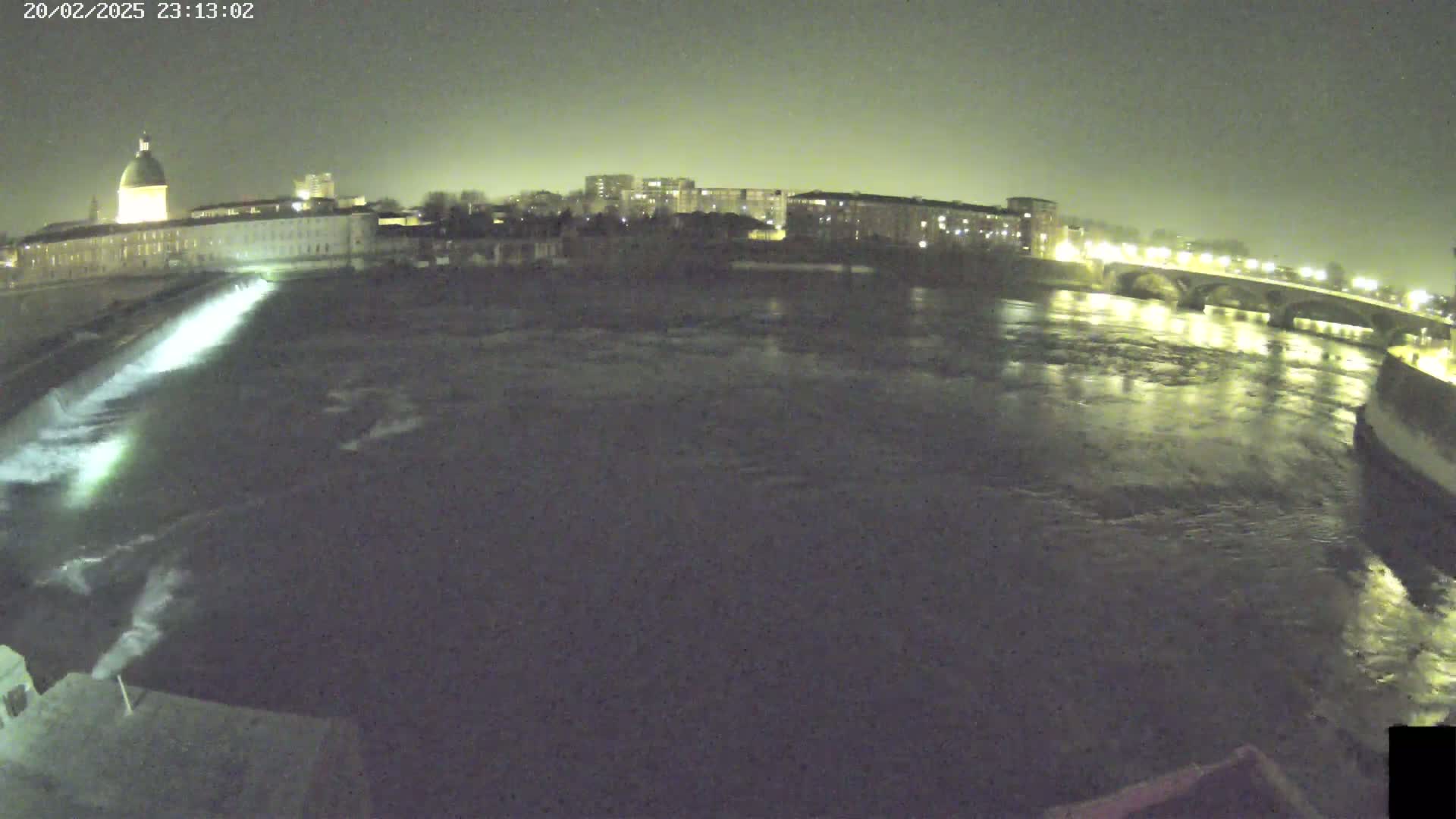 A nighttime view of a river flowing past city buildings and a bridge under a dark sky.