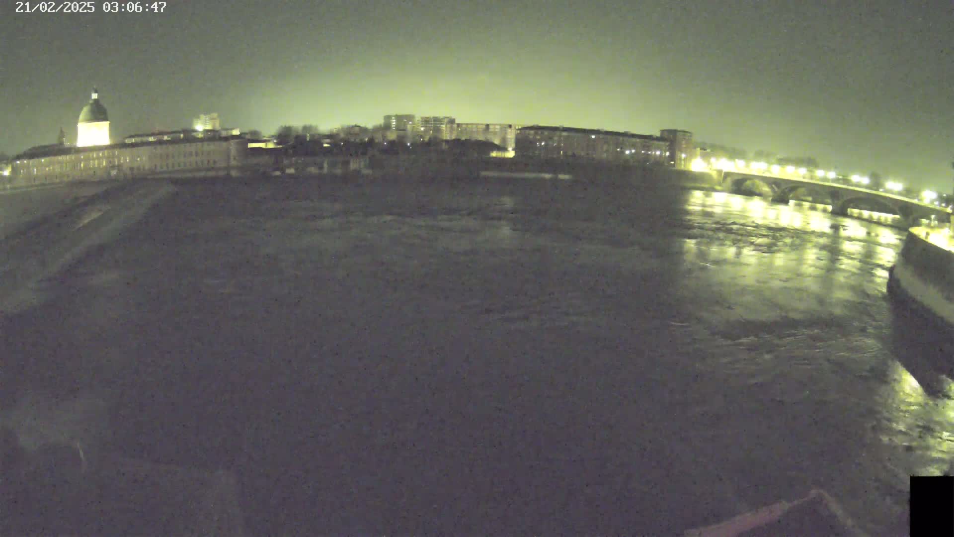 A nighttime view of a river flowing past city buildings and a bridge under a dark sky.