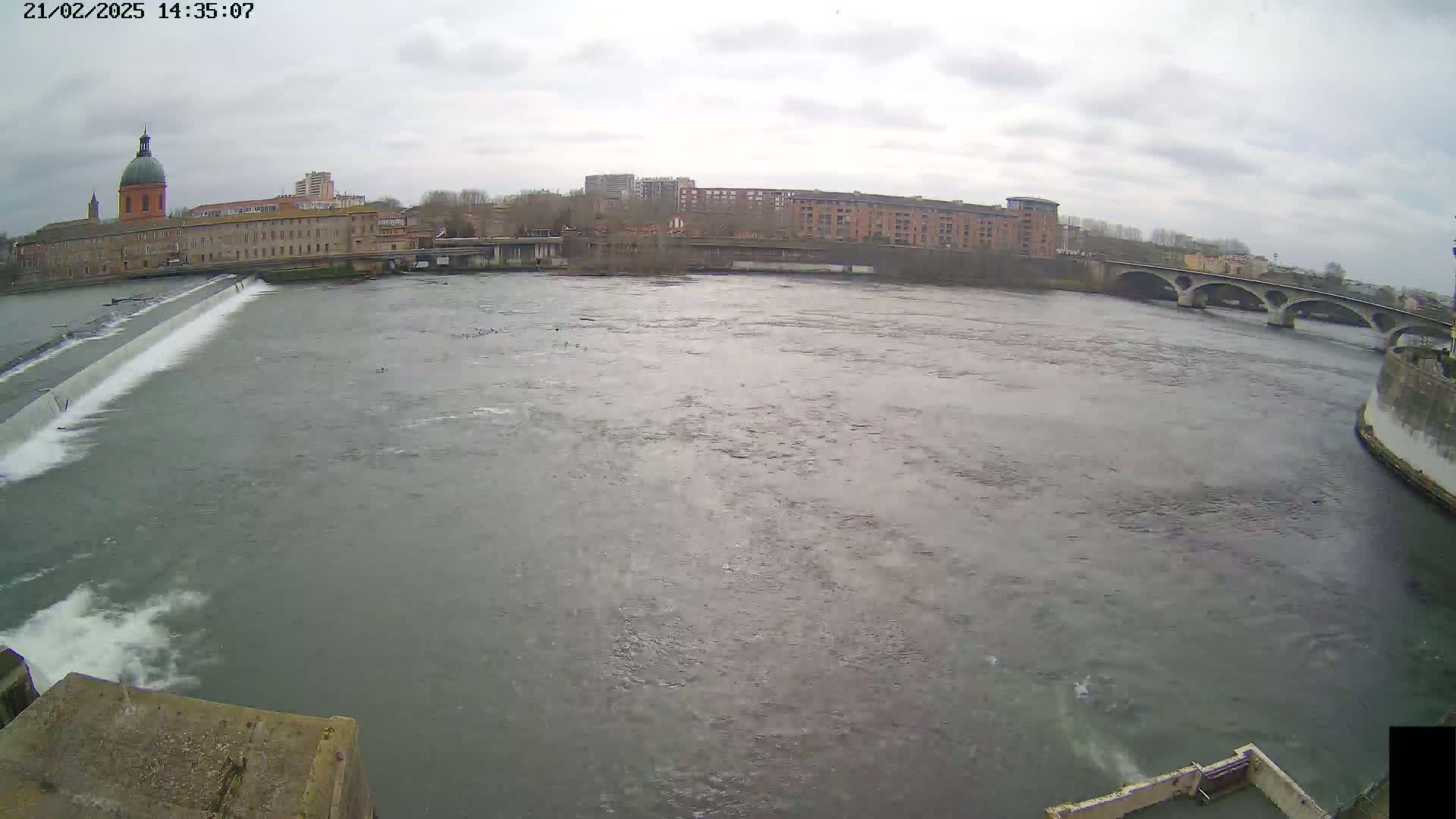 A wide shot of a river flowing past a city under an overcast sky, featuring a dam and a bridge in the distance.