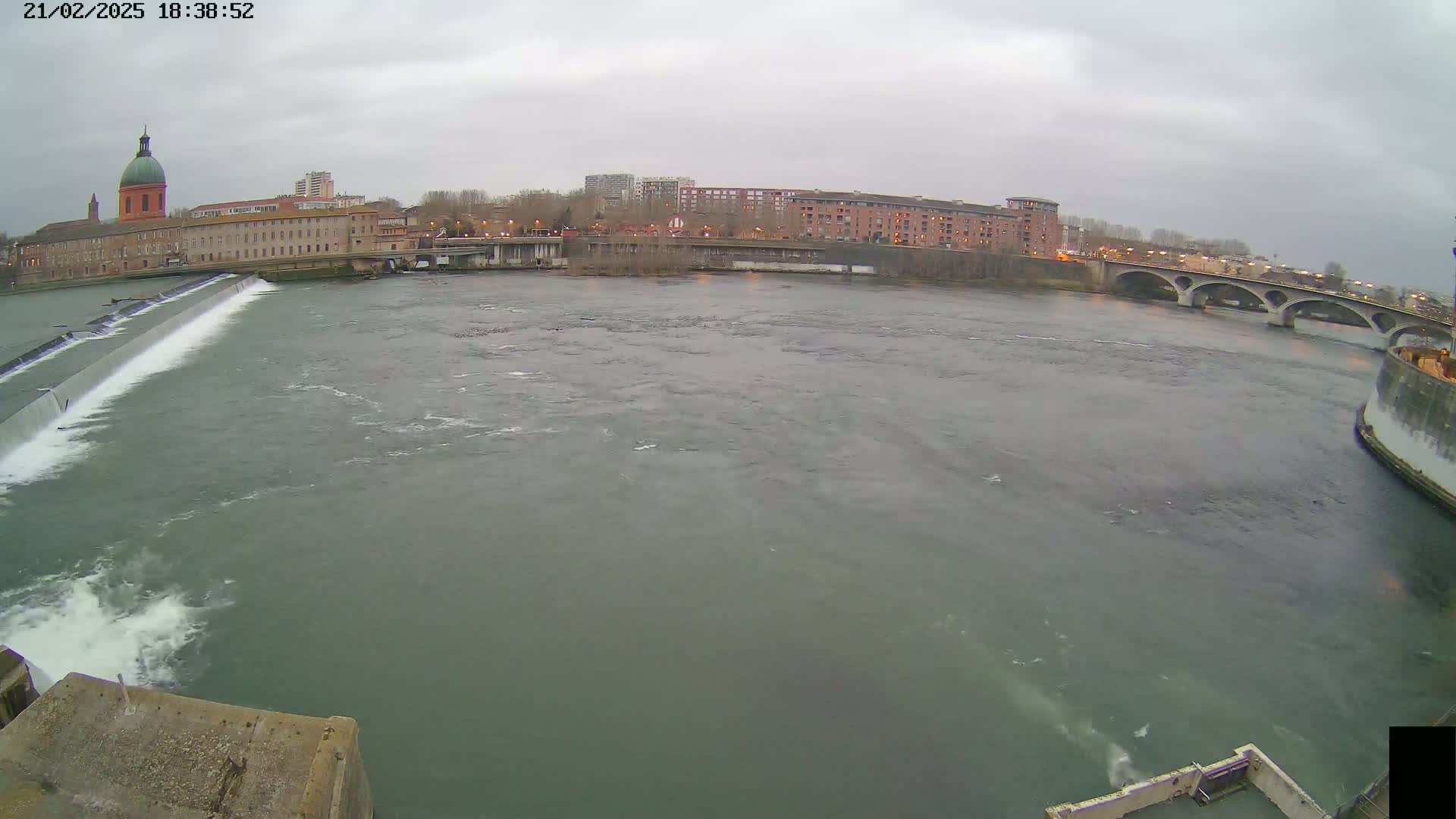 A wide river flows under an overcast sky past a dam, buildings, and a bridge.