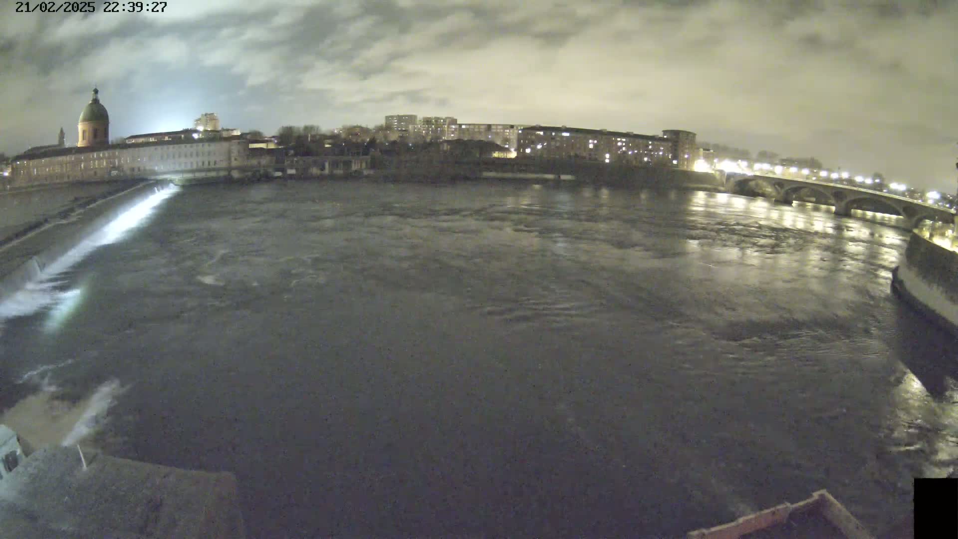 A nighttime view of a river flowing under a bridge past a city with buildings lit up under a cloudy sky.