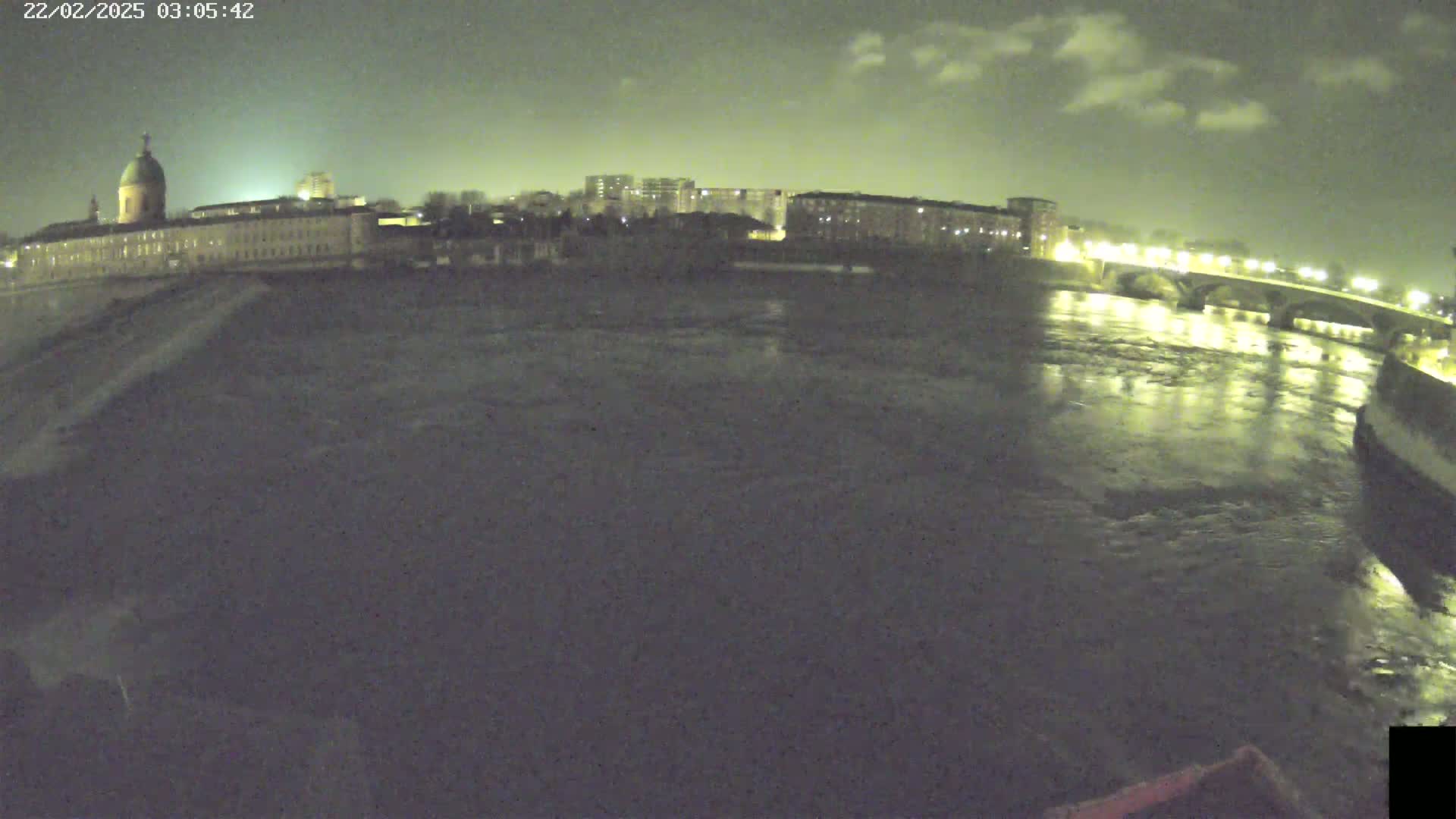 A nighttime view of a river flowing past a city with buildings illuminated, under a partly cloudy sky.