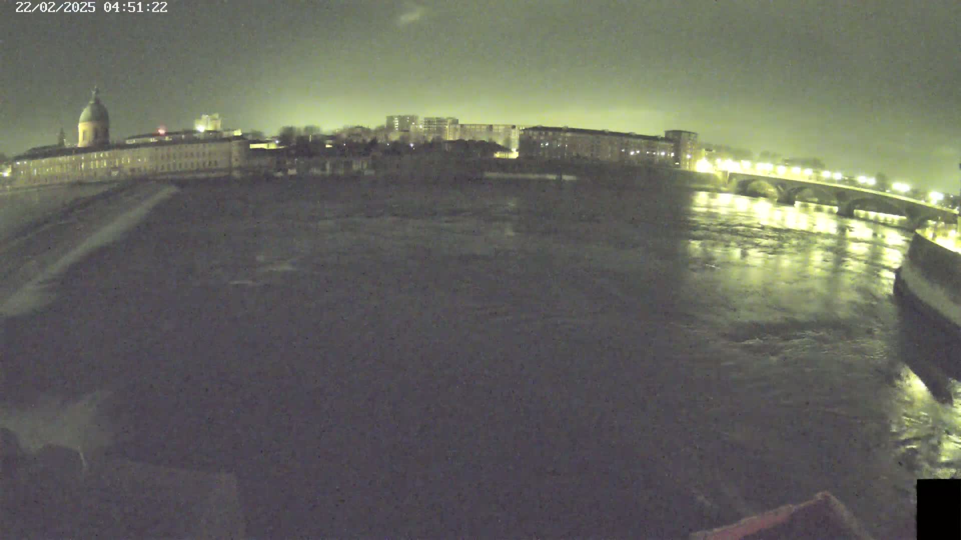 A nighttime view of a river flowing past a city, with buildings and a bridge illuminated under a dark, cloudy sky.