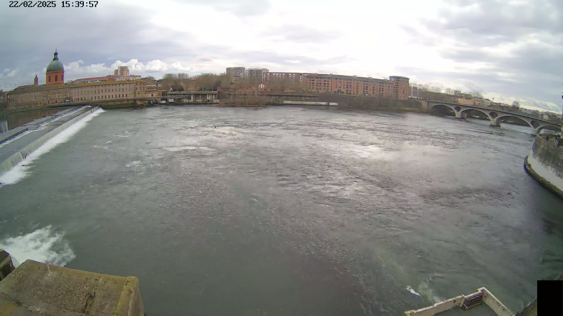 A wide, choppy river flows past a city with buildings and a bridge under an overcast sky.