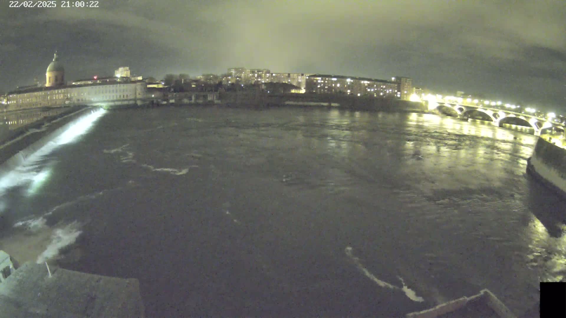 A nighttime view of a river flowing past a city with a dam and bridge under an overcast sky.