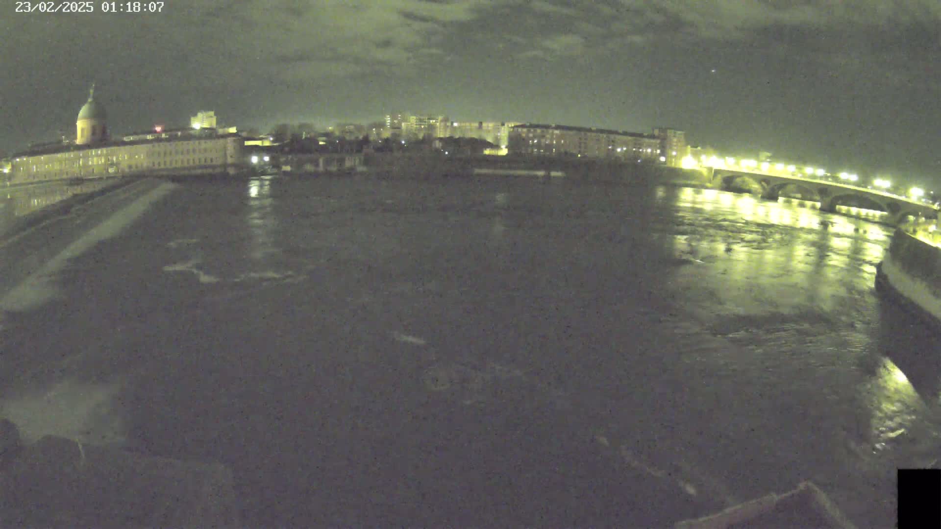 A nighttime view of a river flowing past a city, with a bridge and buildings illuminated under a partly cloudy sky.