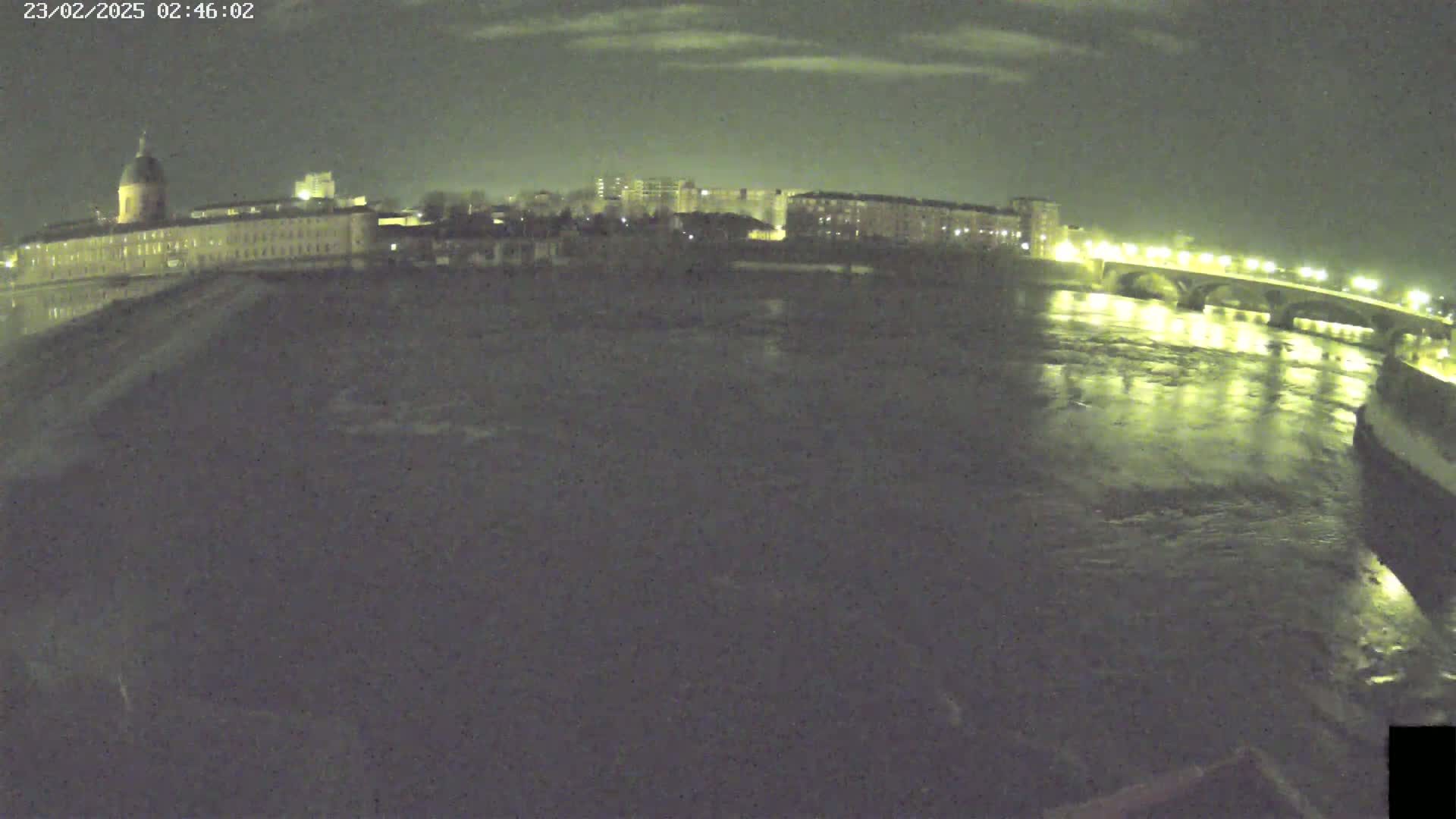 A nighttime view of a river flowing under a lit bridge, with a large building and city skyline visible in the background under a partly cloudy sky.