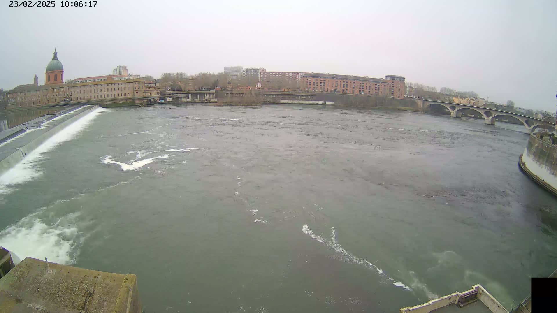 A wide shot shows a river flowing under an arched bridge, past a dam, and alongside buildings on an overcast day.
