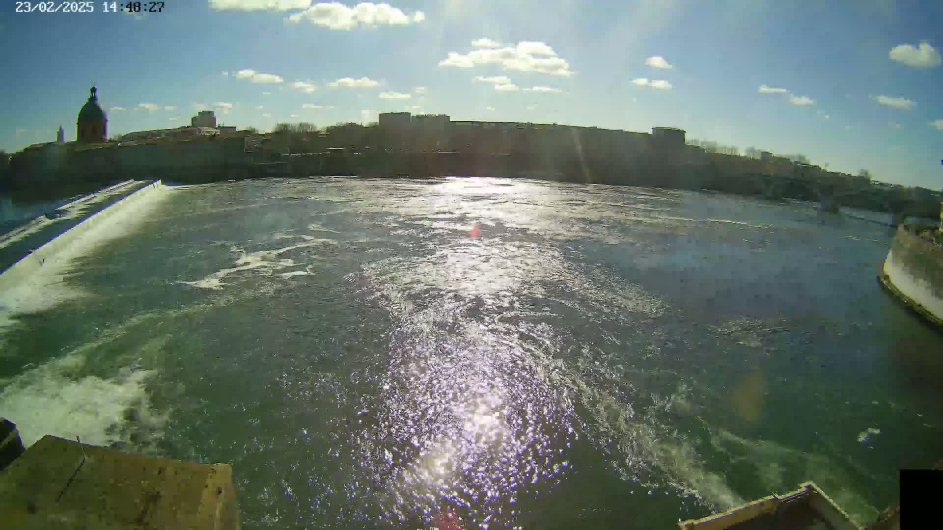 A sunny day shows a river flowing past a dam and city buildings, with some clouds in the sky.