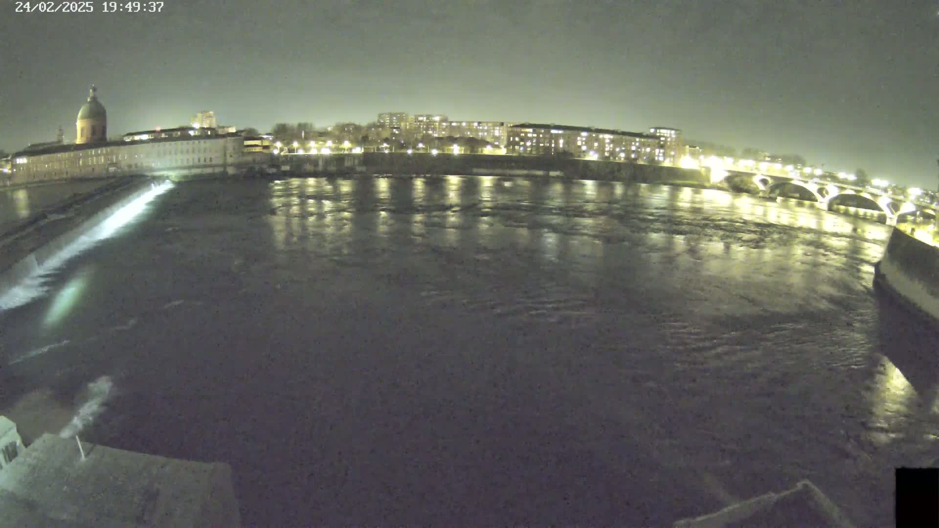 A nighttime view of a river flowing past city buildings and a bridge under a dark sky.