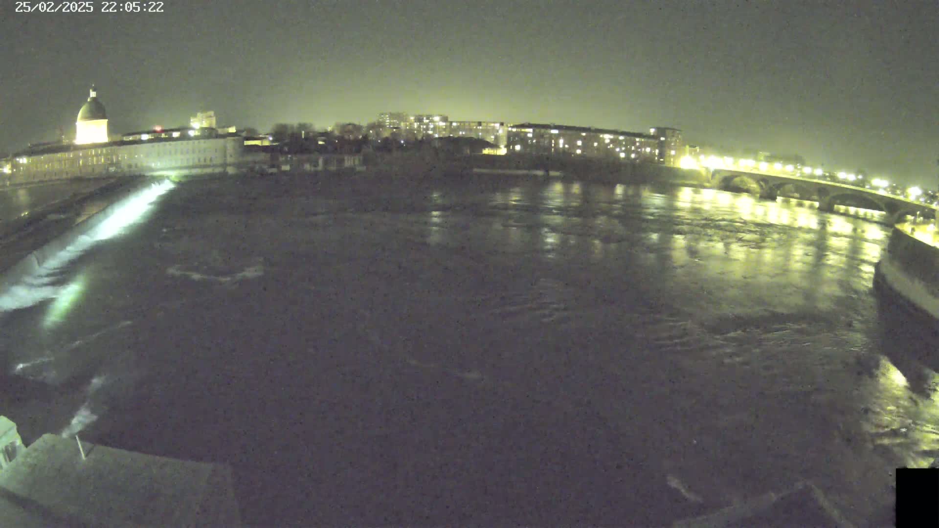 A nighttime view of a river flowing past city buildings and a bridge, with a dam visible in the foreground.