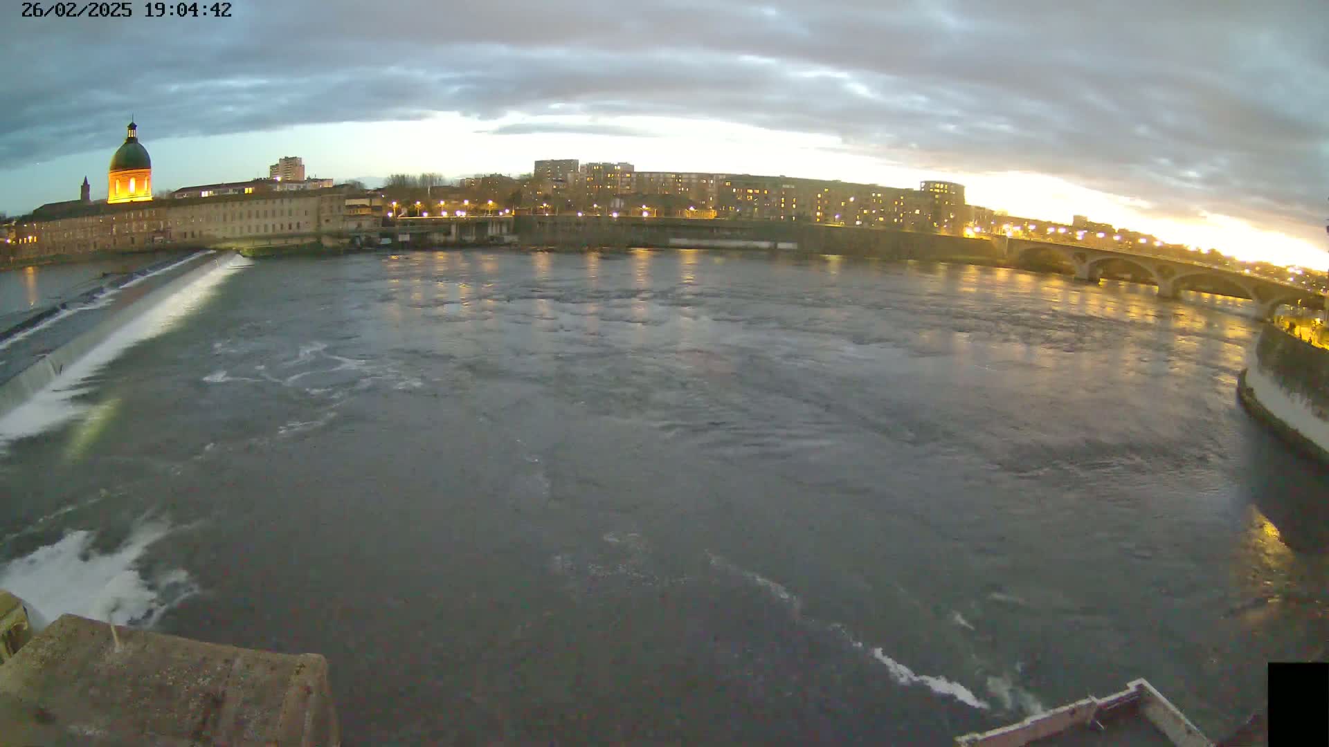 A wide shot shows a river flowing past a city at twilight under a cloudy sky, with a dam visible in the foreground.