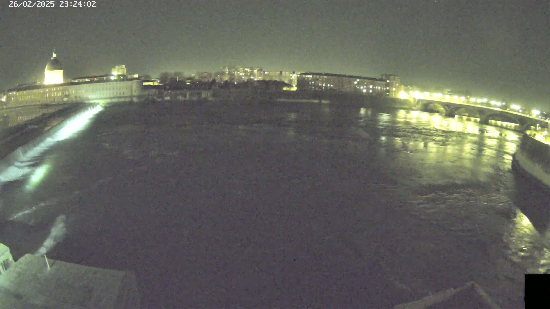 A nighttime view of a river flowing past a city, with a dam releasing water and a bridge illuminated in the distance.