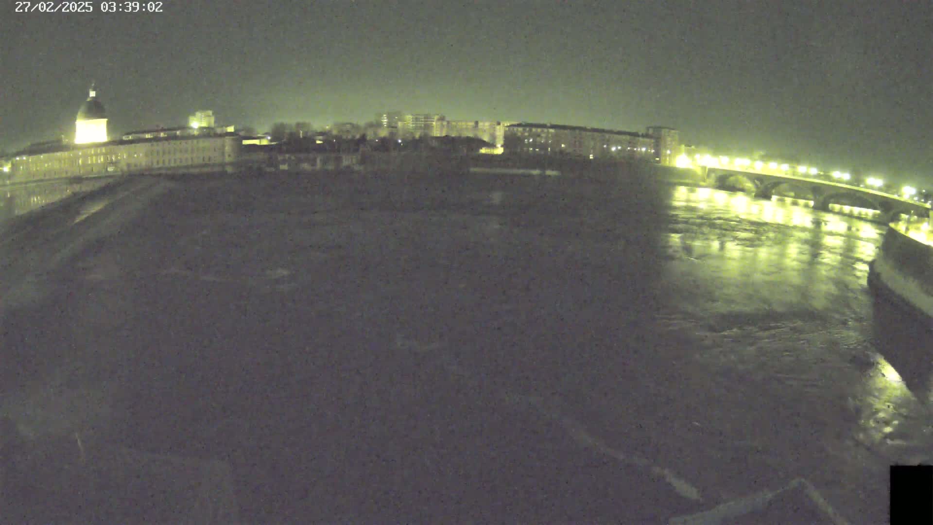 A nighttime, wide shot shows a river flowing under a bridge, with city buildings and lights visible on both sides of the riverbank under a dark sky.
