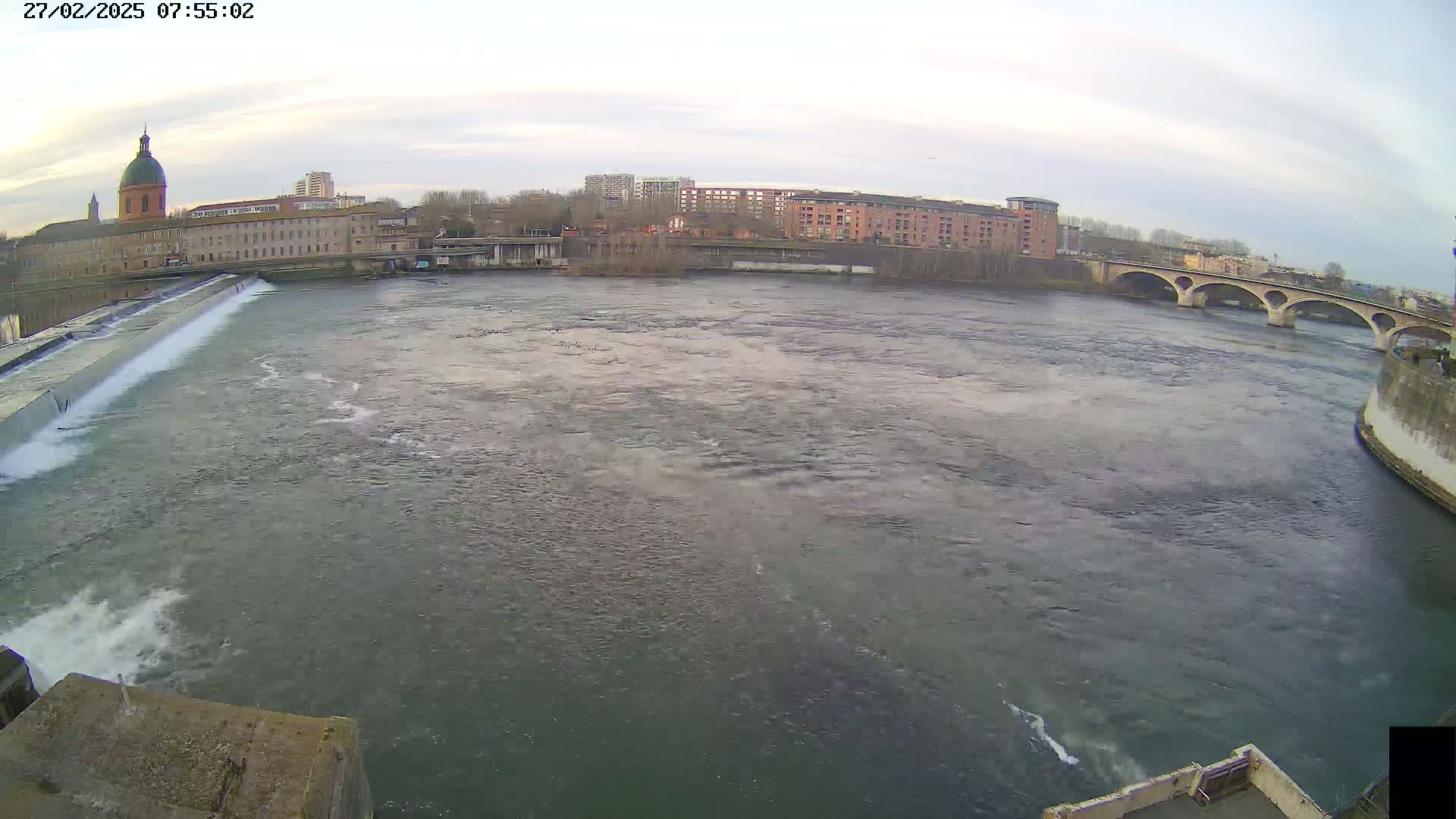 A wide shot of a river flowing past a city under an overcast sky, featuring a dam, buildings, and a bridge.