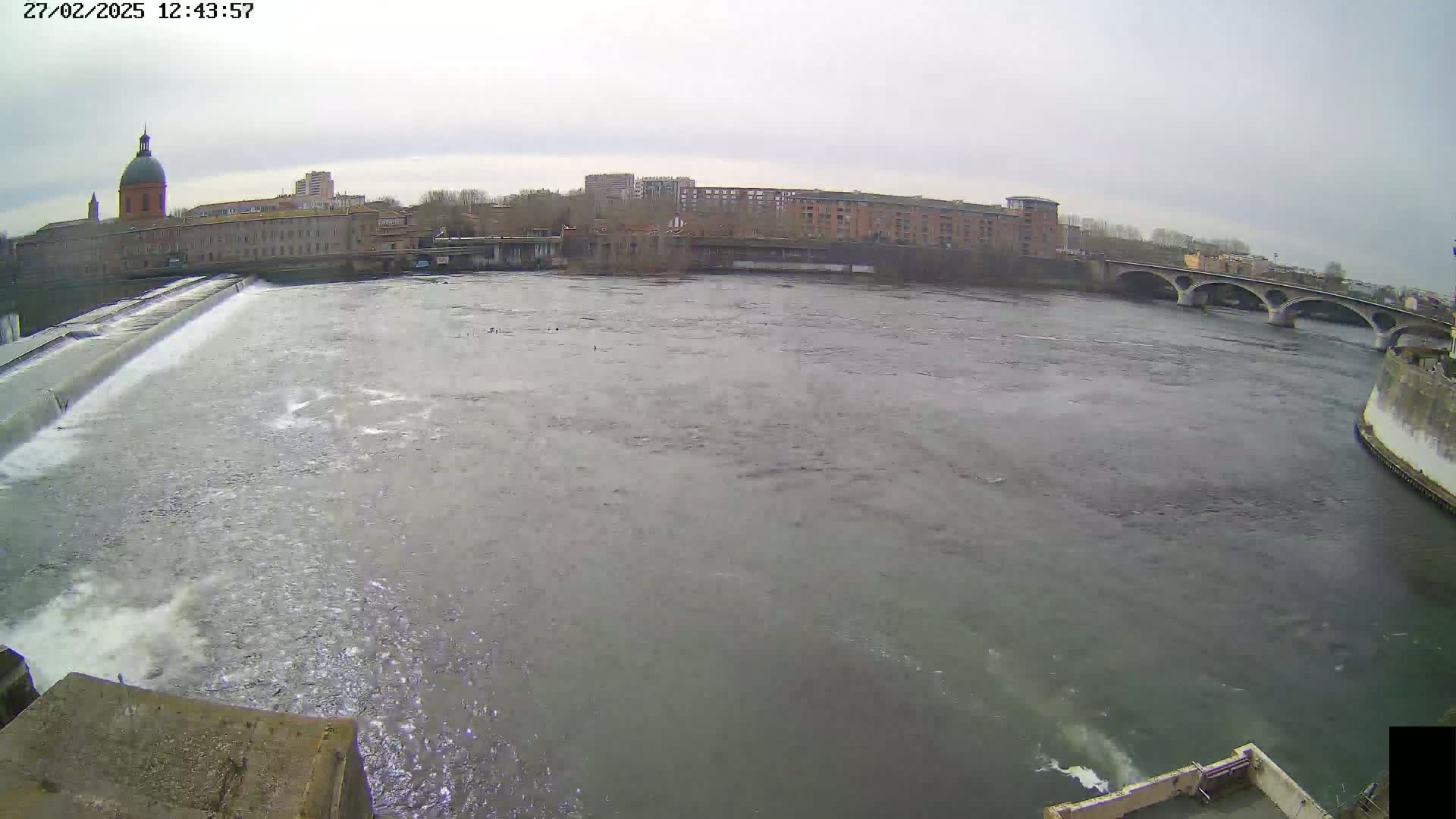 A wide shot of a river flowing under a bridge past a city with overcast skies.