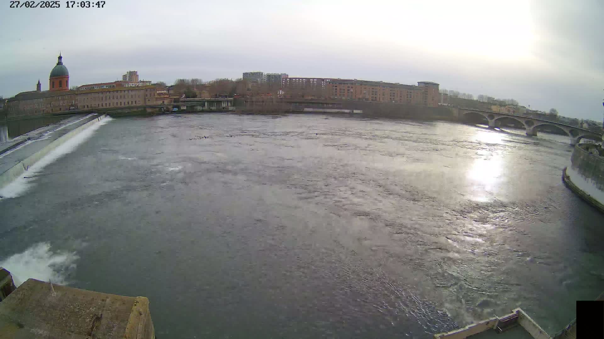 A wide shot of a river flowing past a city with a dam and bridge under an overcast sky.