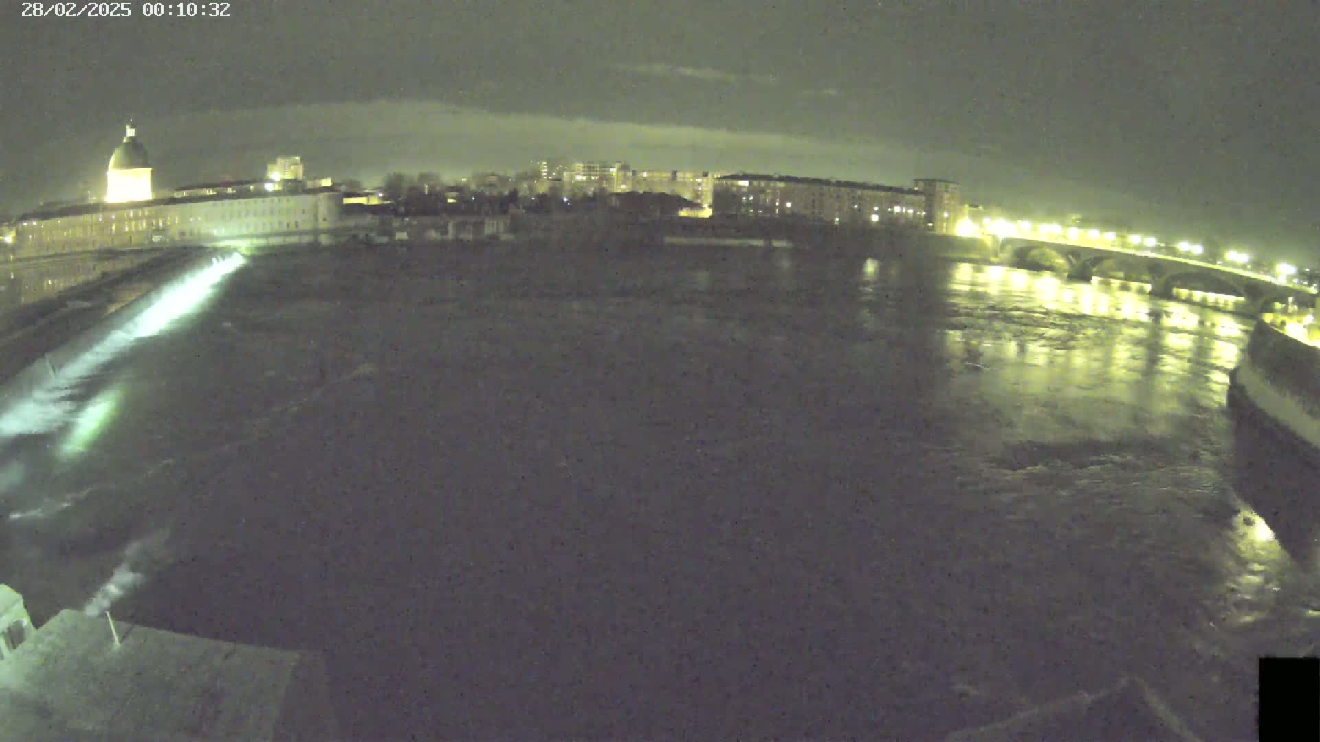 A nighttime view of a river flowing past a city, with a dam and bridge illuminated.