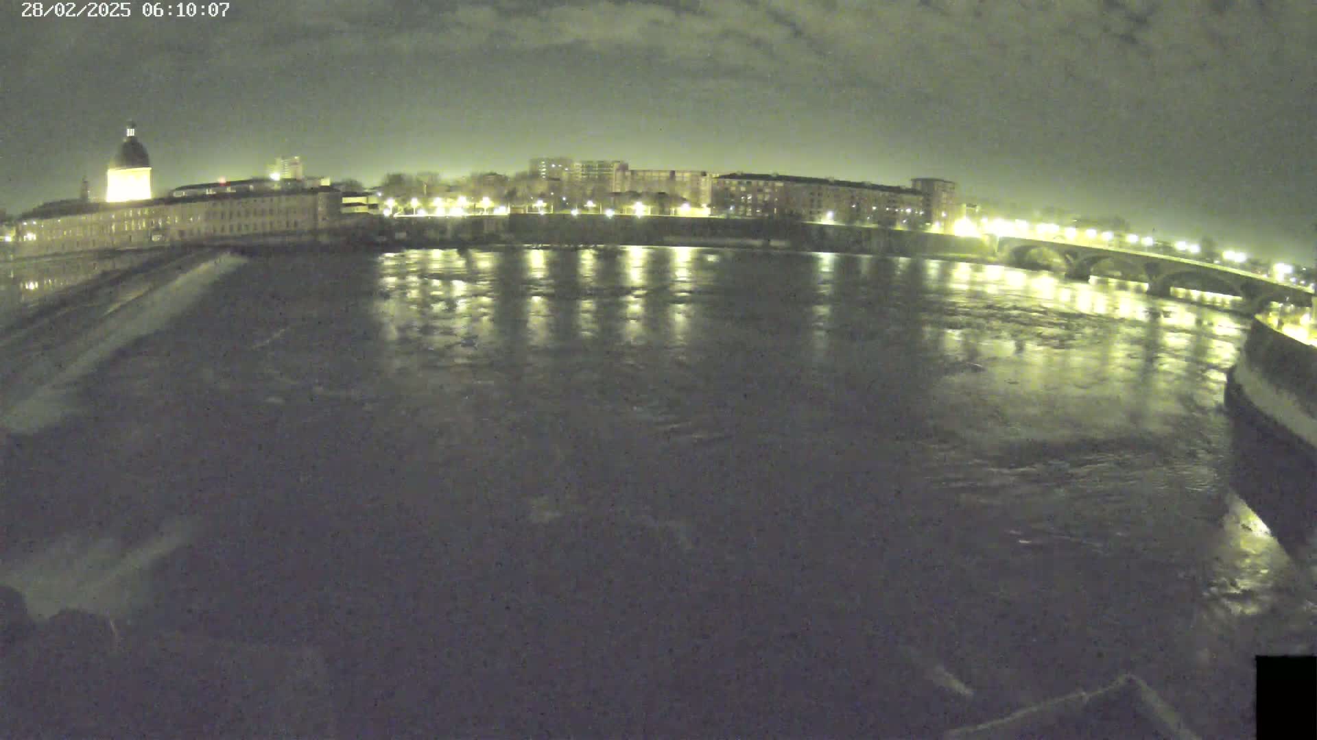 A nighttime view of a river flowing under a bridge, past buildings with lights illuminating the water and the cityscape under a cloudy sky.