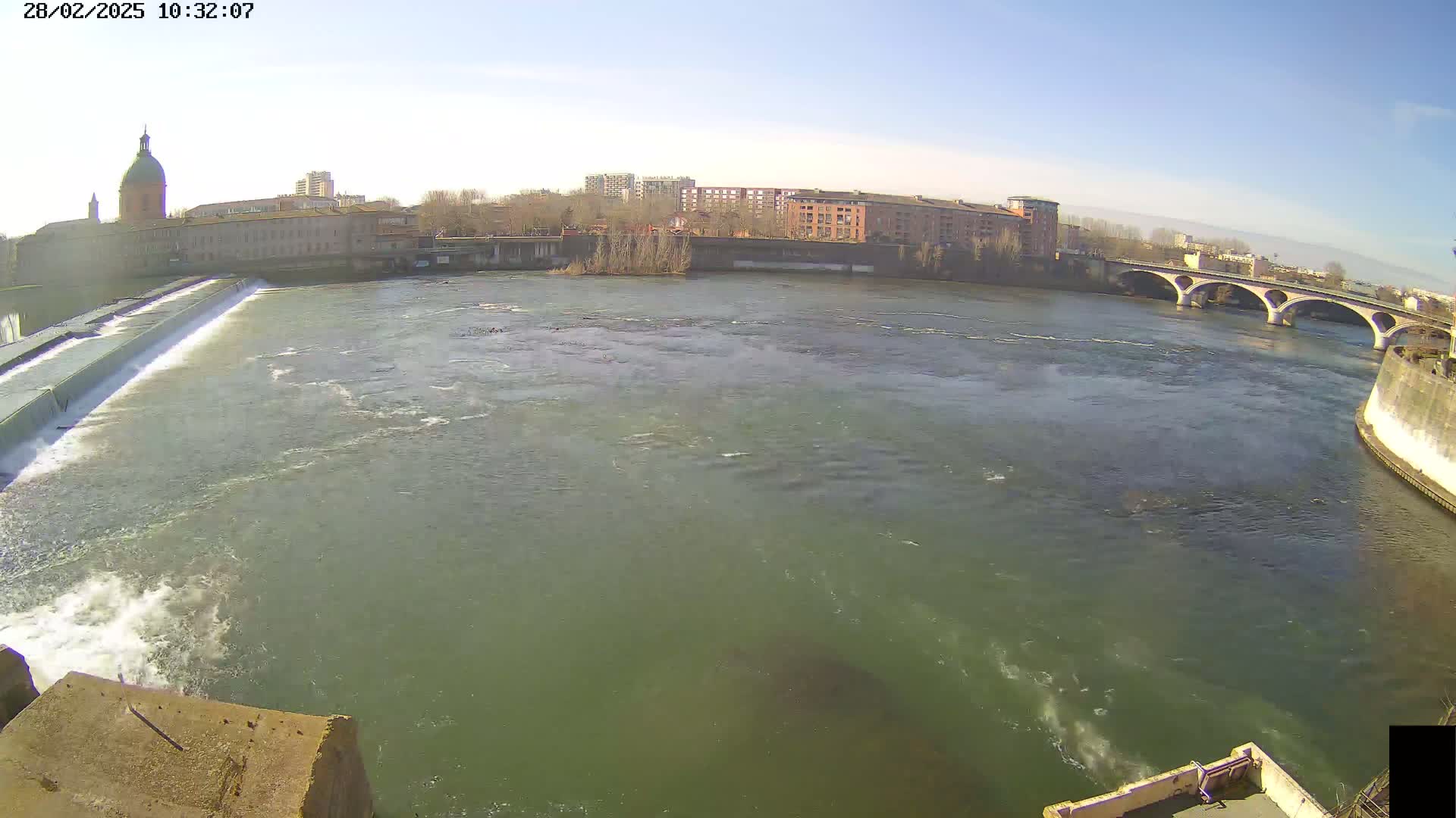 A wide shot shows a river flowing past a dam and under a bridge on a sunny day, with buildings visible along the riverbank.