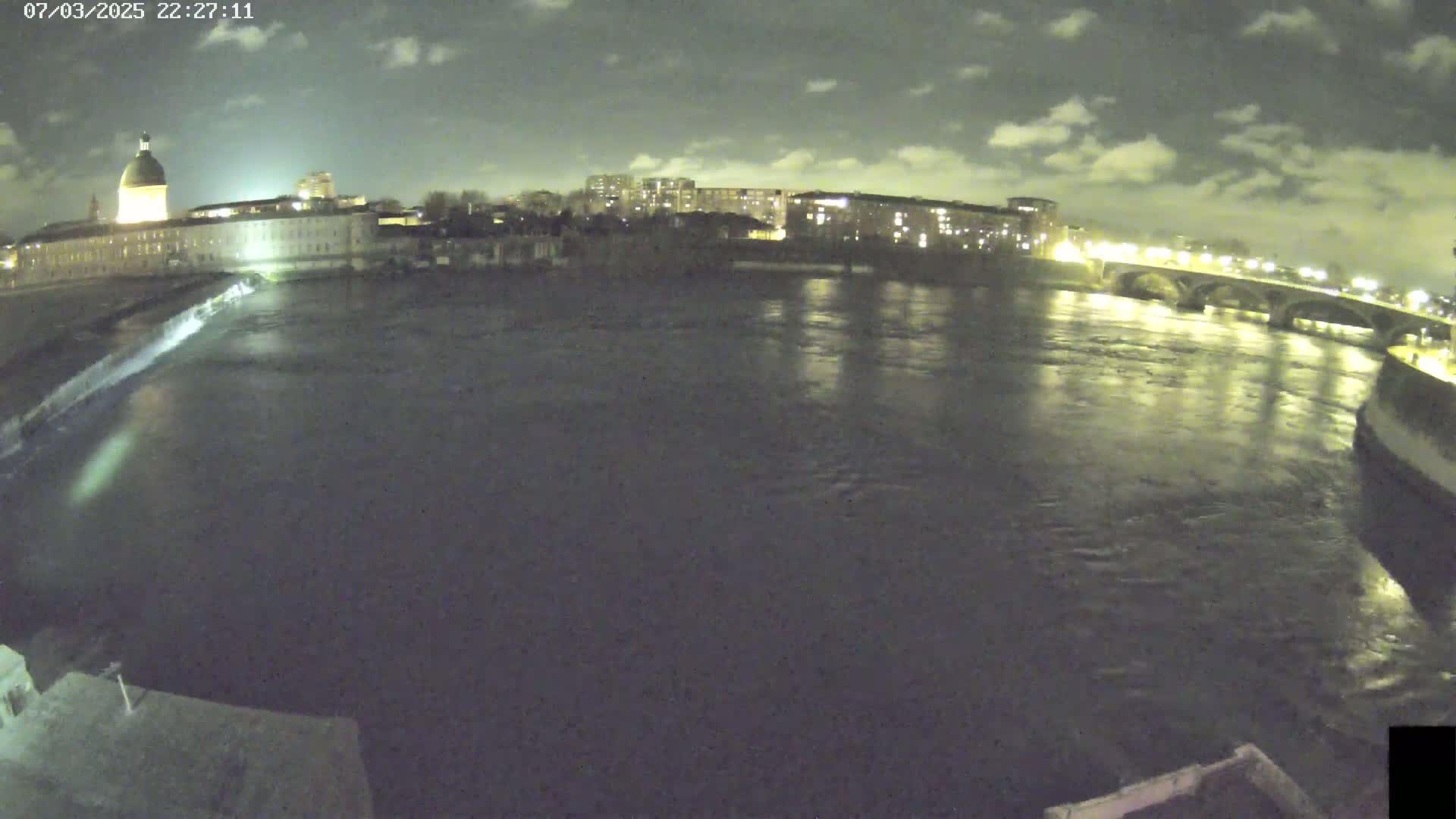 A nighttime view of a river flowing past city buildings and a bridge under a cloudy sky.
