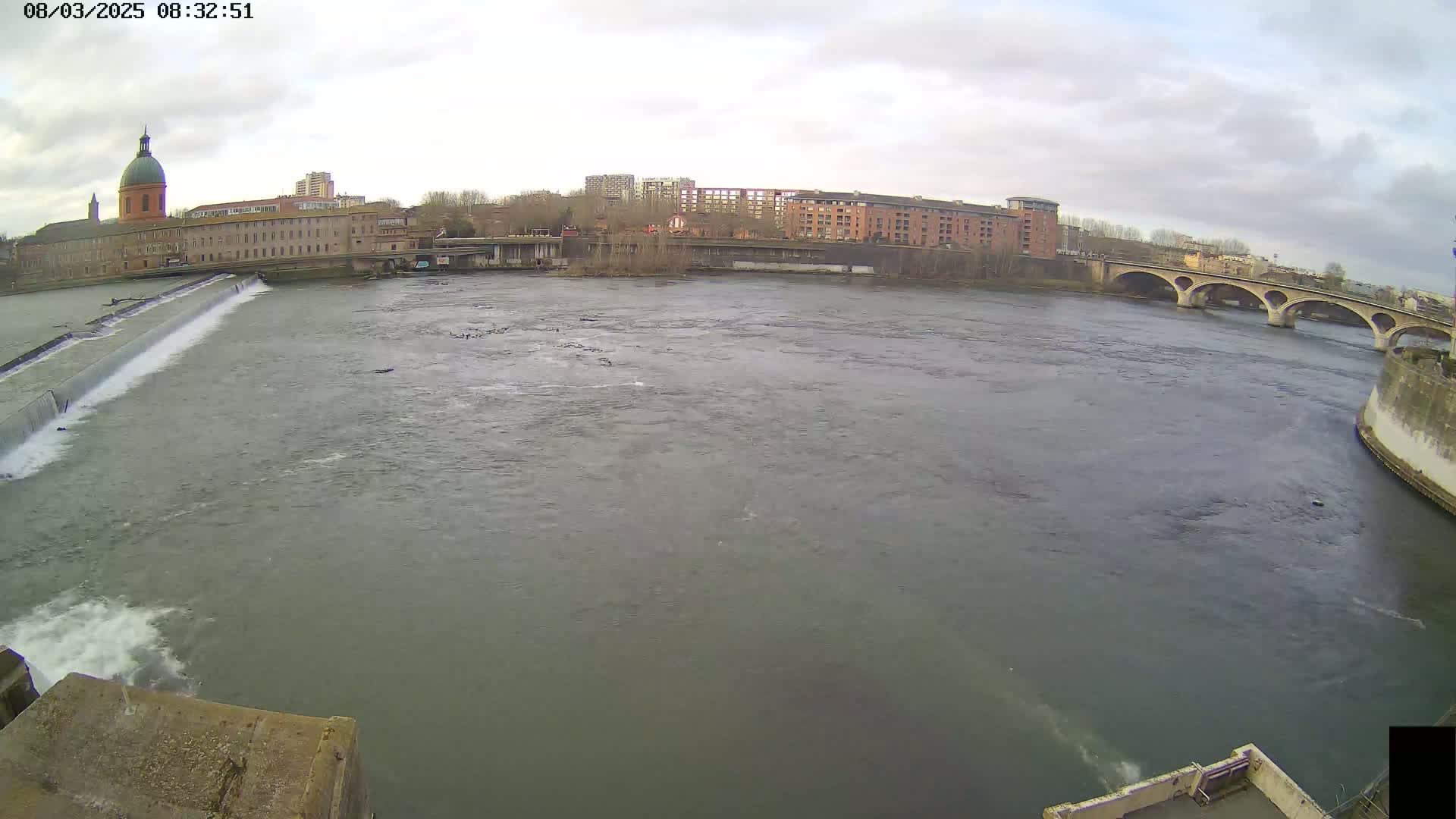 A wide shot shows a river flowing under an arched bridge, past a dam and buildings on an overcast day.