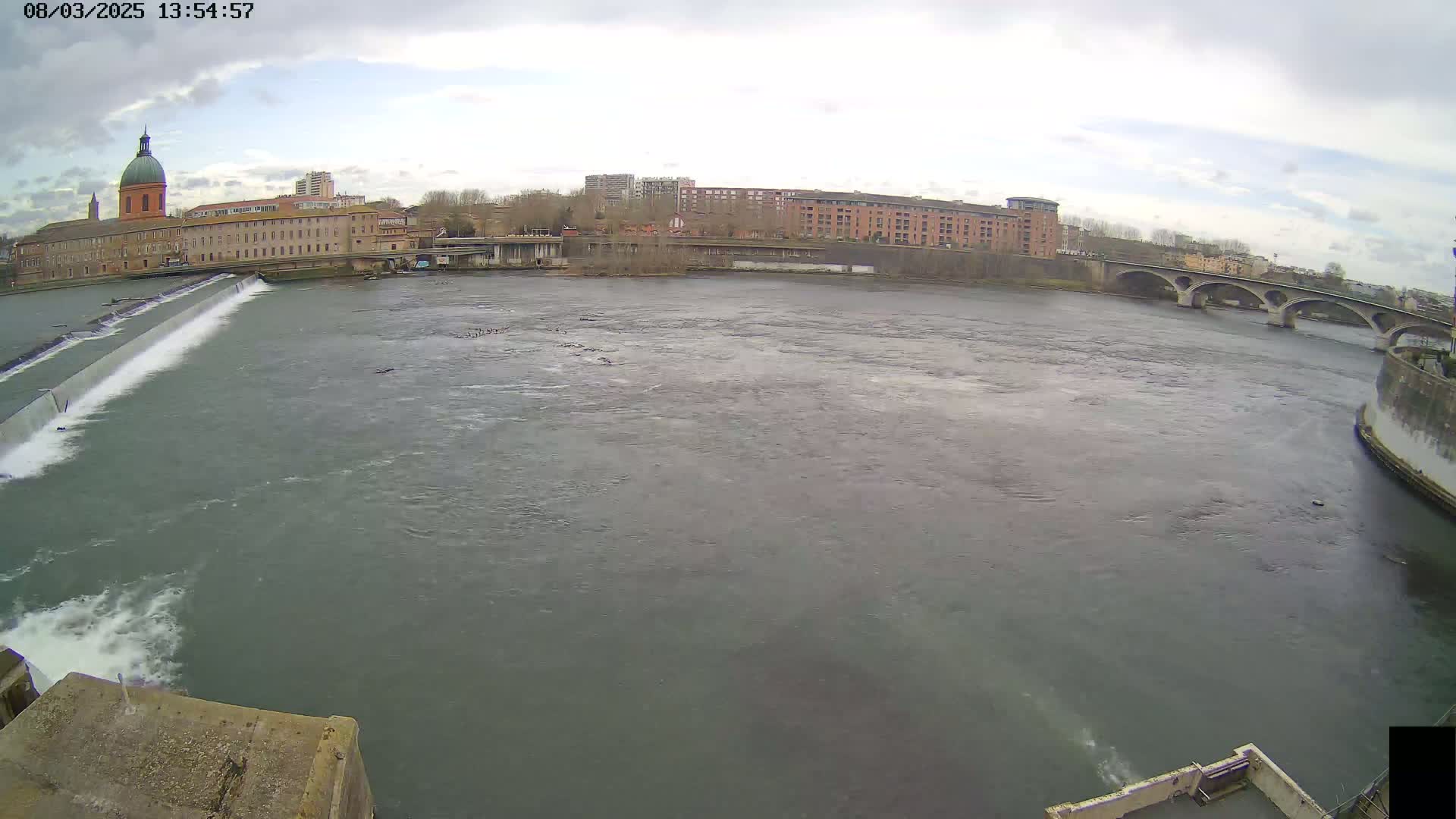 A wide shot of a river flowing past a city under an overcast sky, featuring a dam, buildings, and a bridge in the distance.